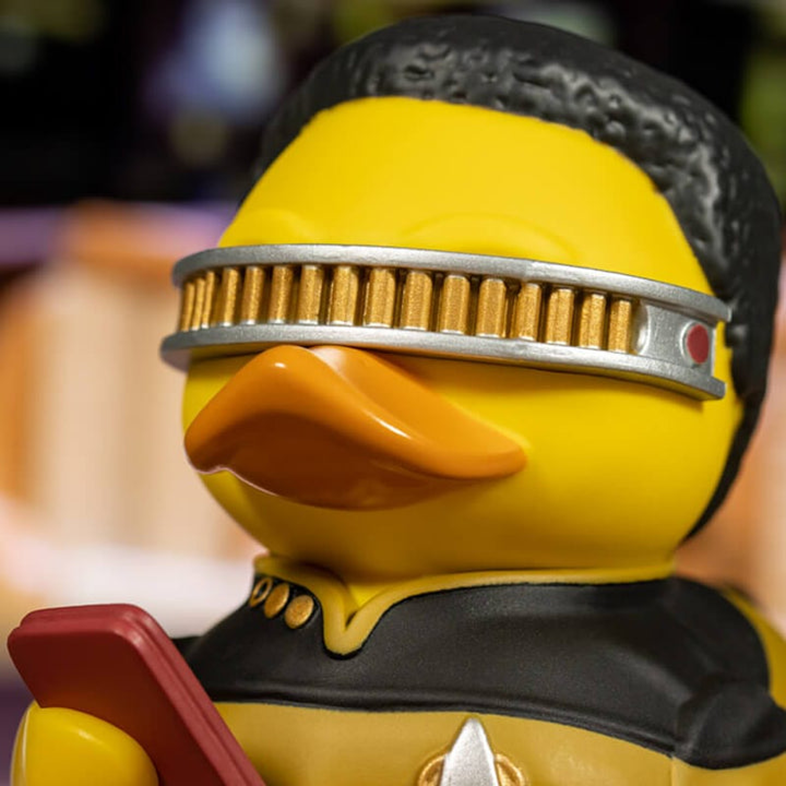 Egy sárga kacsa játék Star Trek egyenruhában és napellenzővel, egy piros eszközt tartva.