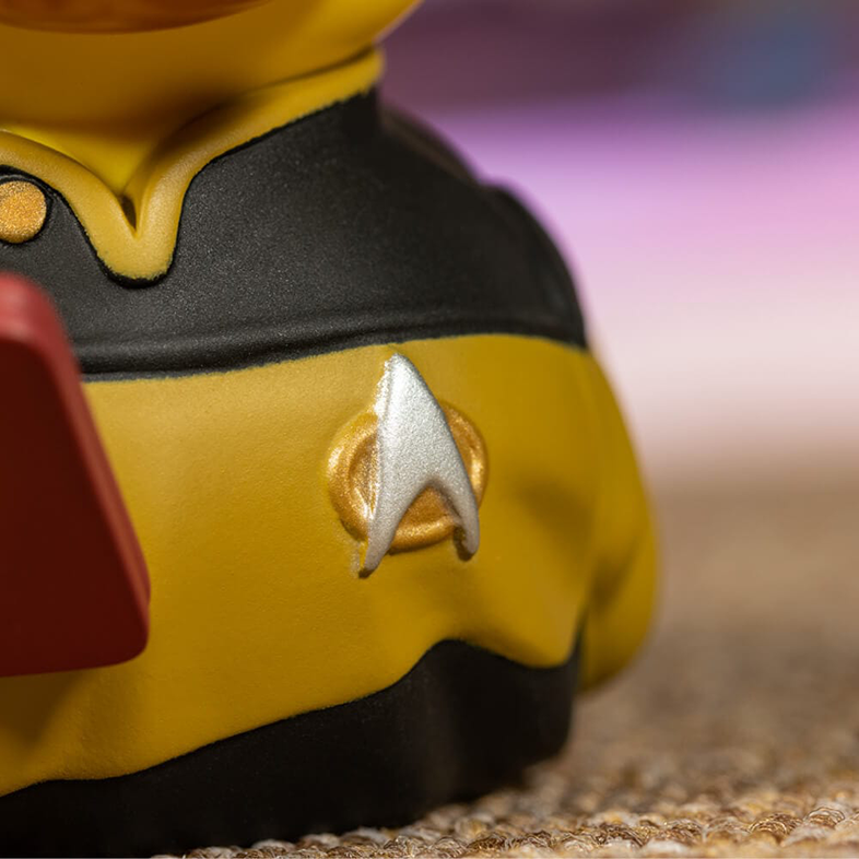 Egy sárga figura vállának közelképe Star Trek jelvénnyel. A figura sárga egyenruhát visel. Homályos háttér.
