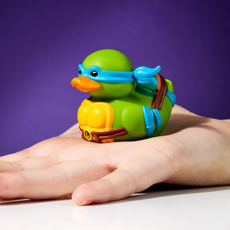 Egy zöld gumikacsa, Teenage Mutant Ninja Turtles-ként tervezve, egy kézen ül. Lila háttér.