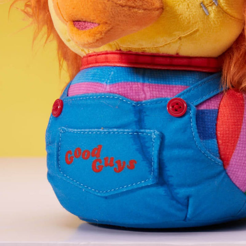 Egy Chucky baba kék overállal és 'Good Guys' felirattal a zsebén. A háttér sárga.