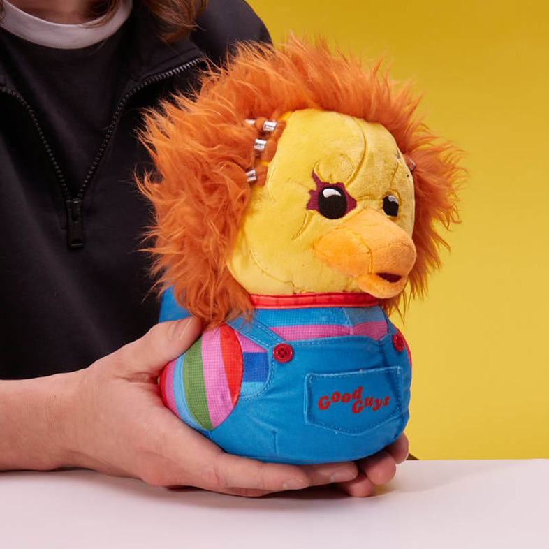 Egy Chucky baba, egy plüssjátékot tart egy személy. Sárga és narancssárga, kék overállal.