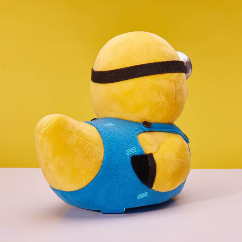 Egy Minion plüssfigura kék overálban, fehér felületen ül, sárga háttérrel.