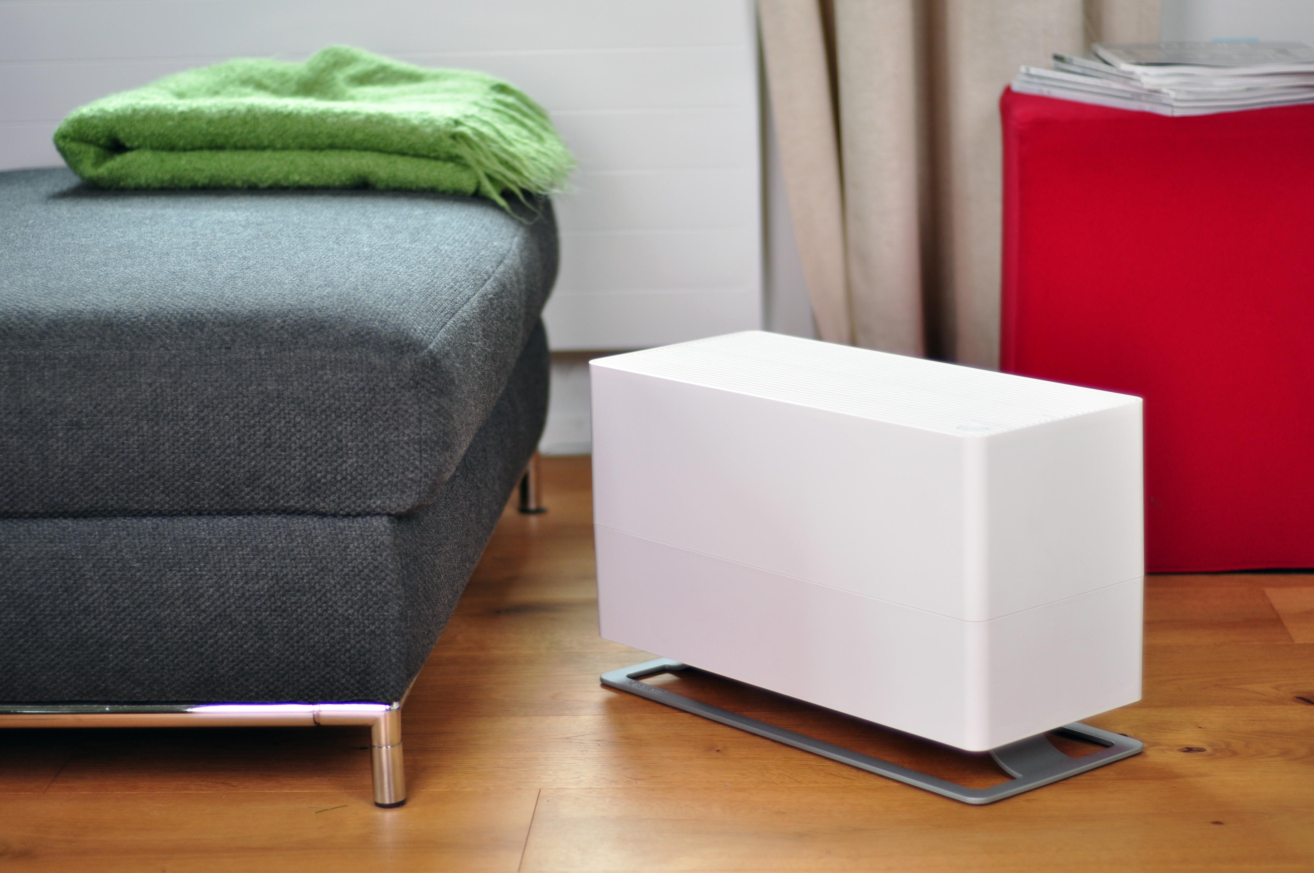 Weißer Luftreiniger neben einem grauen Sofa und einem roten Ottoman.