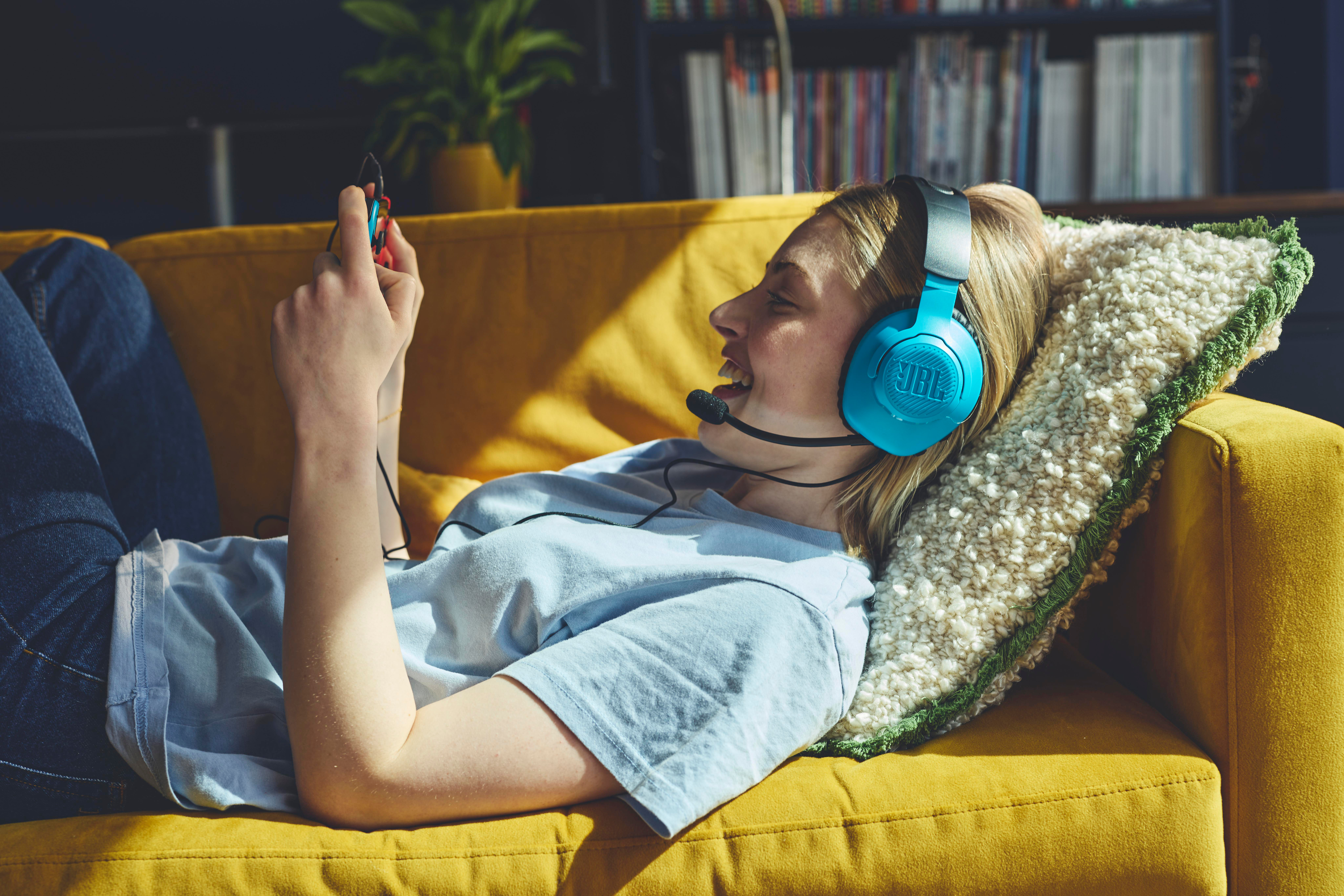 Person auf gelbem Sofa mit blauen Kopfhörern spielt Handheld-Spiel, lächelt. Bücherregal im Hintergrund.