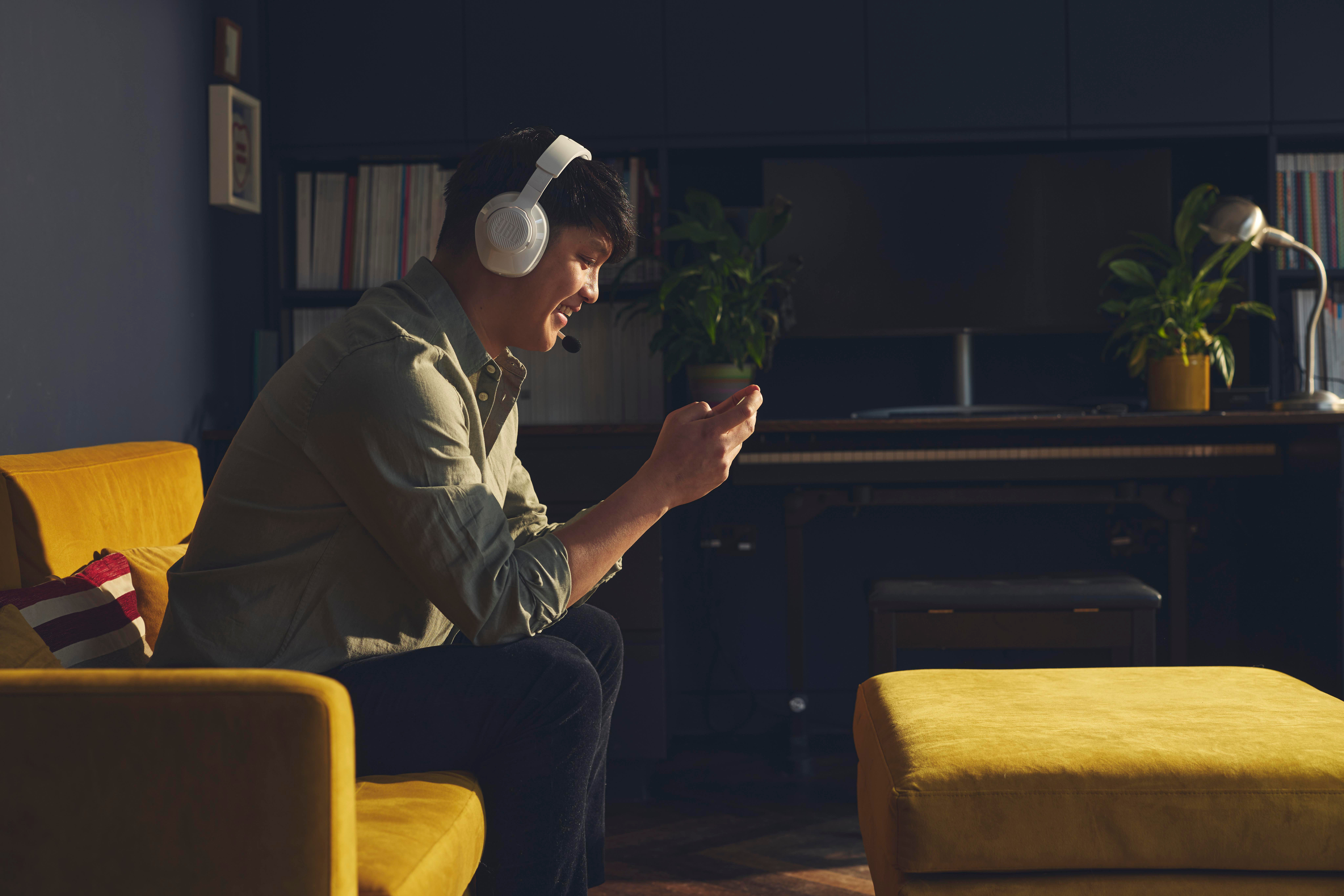 Ein Mann sitzt auf einem gelben Sofa, trägt ein Headset und hält ein Telefon. Im Hintergrund befinden sich ein Klavier und ein Bücherregal.