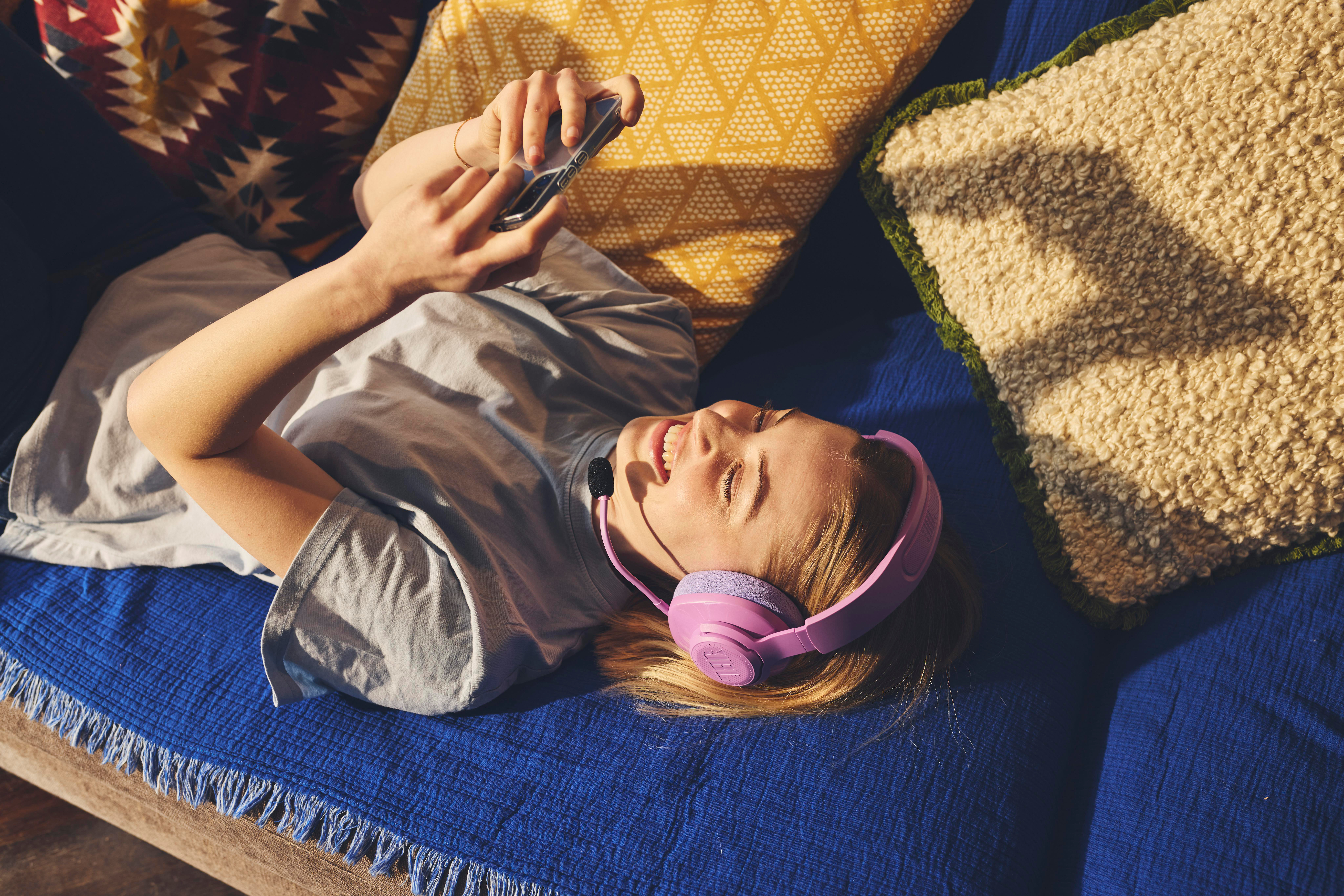 Eine Frau liegt auf einer blauen Couch und trägt rosa Kopfhörer. Sie schaut auf ihr Handy.