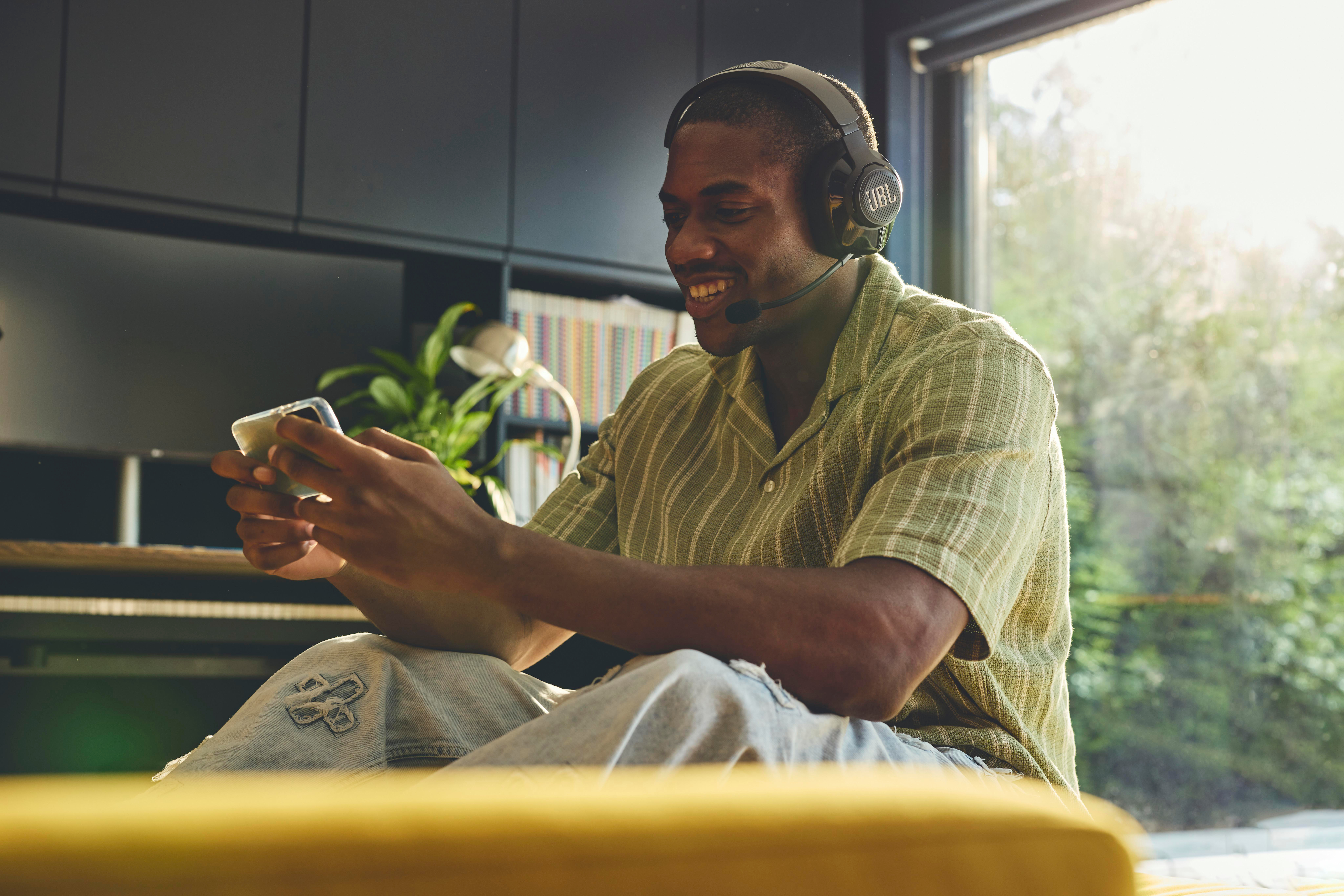 Ein Mann mit Kopfhörern lächelt, während er ein Telefon benutzt, auf Couch sitzt.