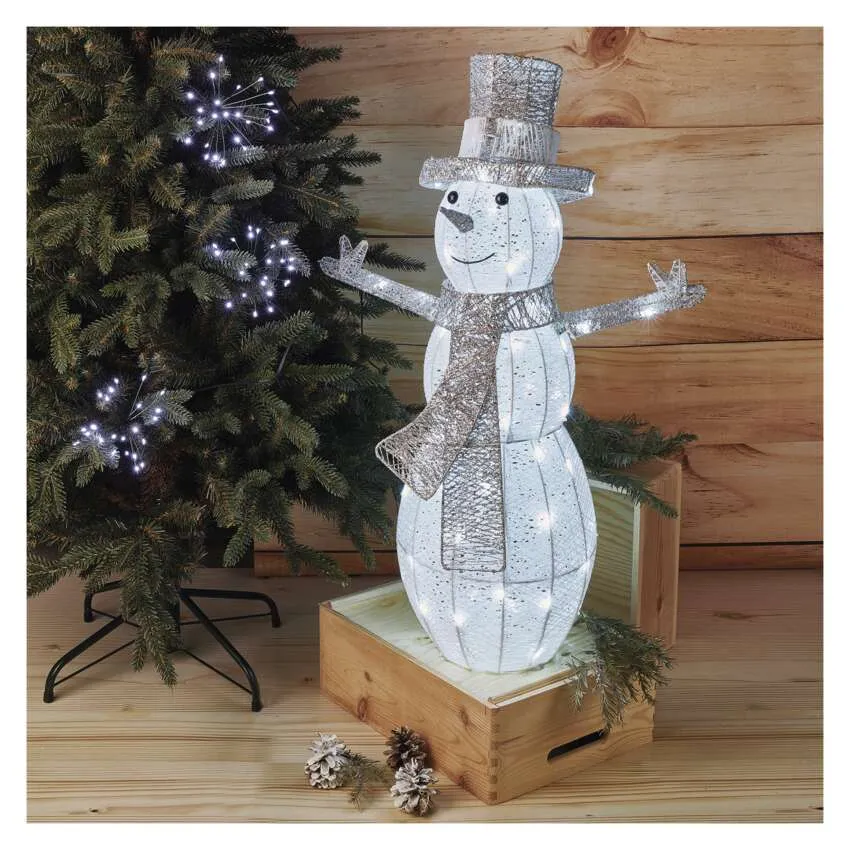 Egy hóember-díszítés lámpákból, egy fadobozon állva egy karácsonyfa közelében.