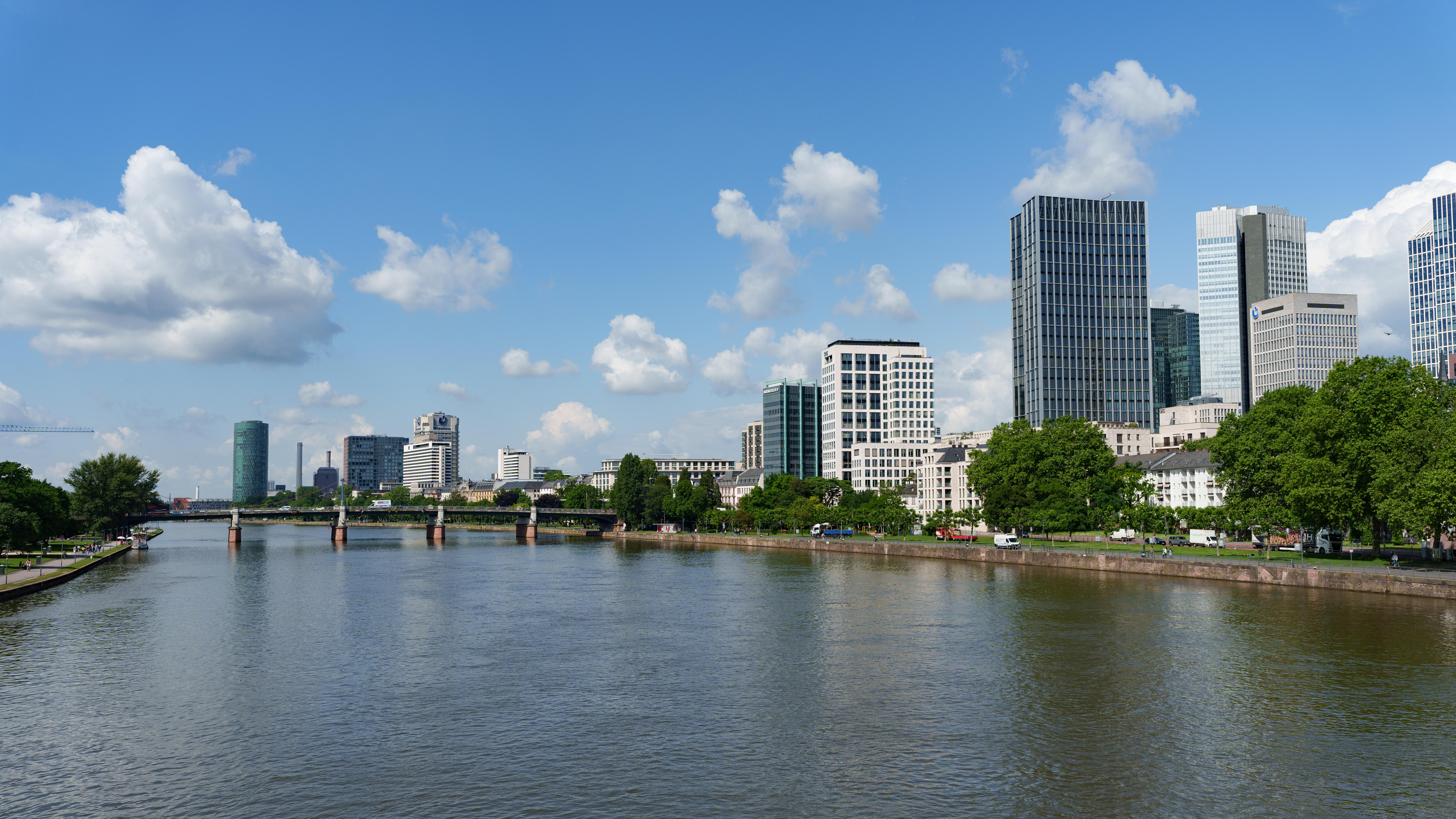 Eine Stadtlandschaft mit Fluss, hohen Gebäuden und Bäumen vor blauem Himmel.