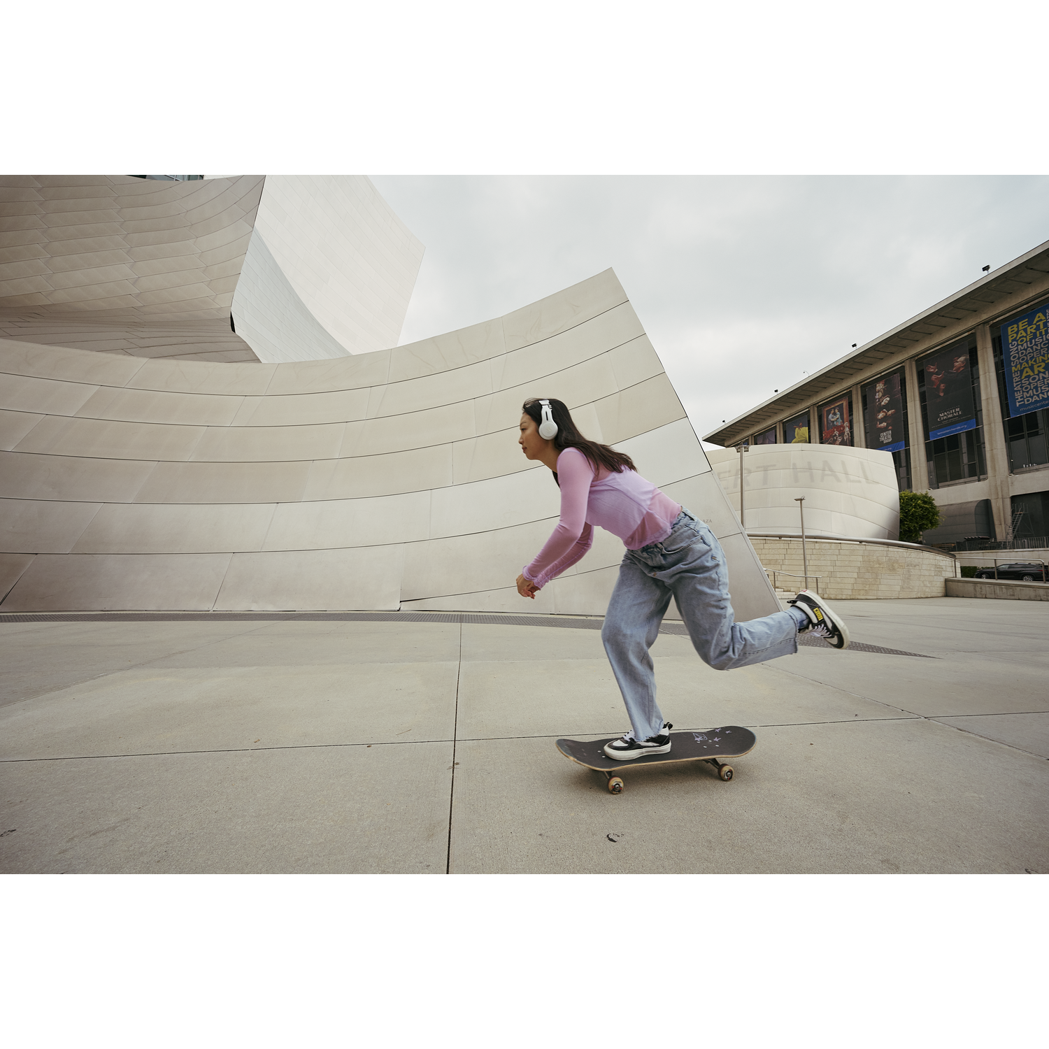 Woman skates on a skateboard in front of a large modern building wearing headphones.