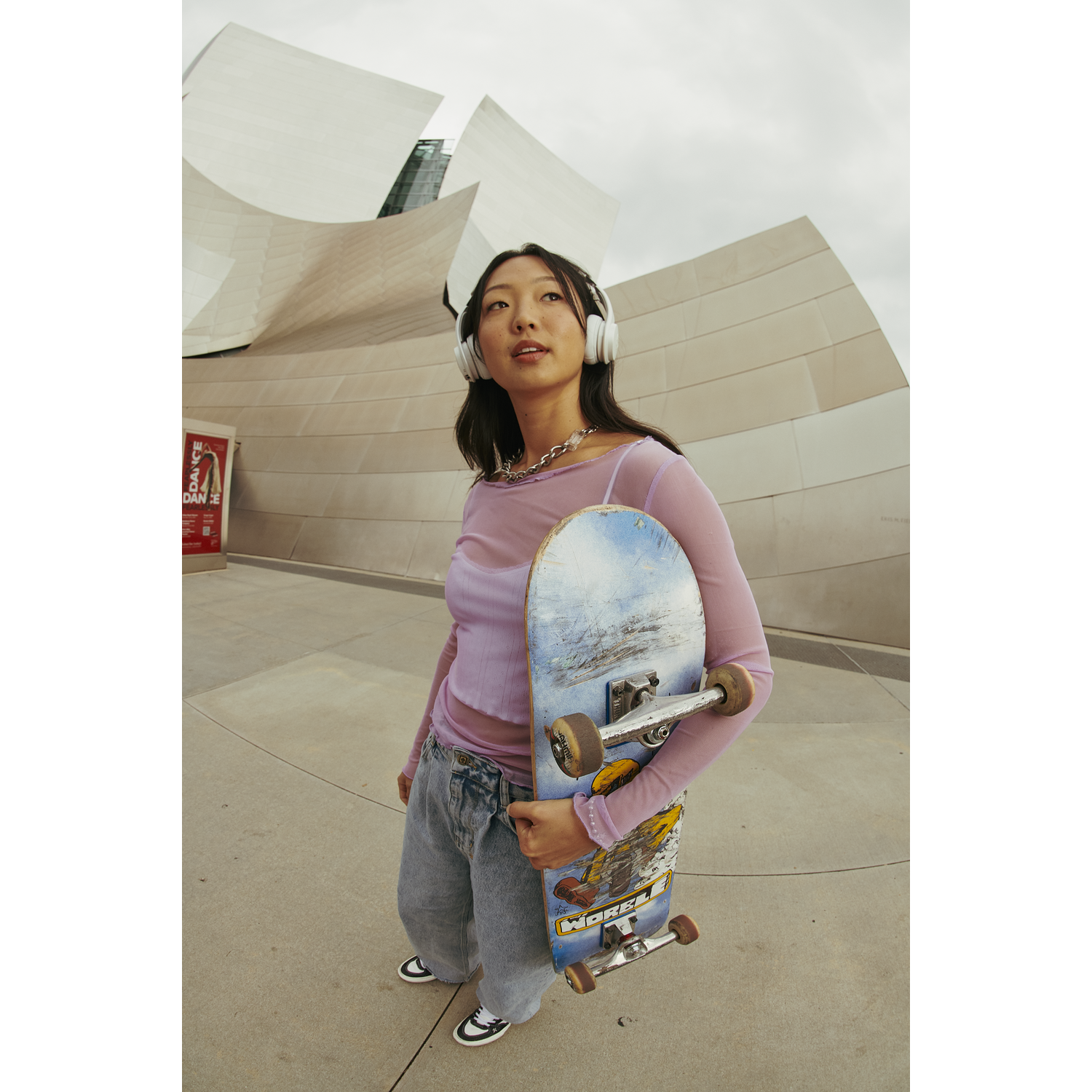Young woman with headphones, skateboard, wearing pink top, jeans, and sneakers, stands in front of a modern building.