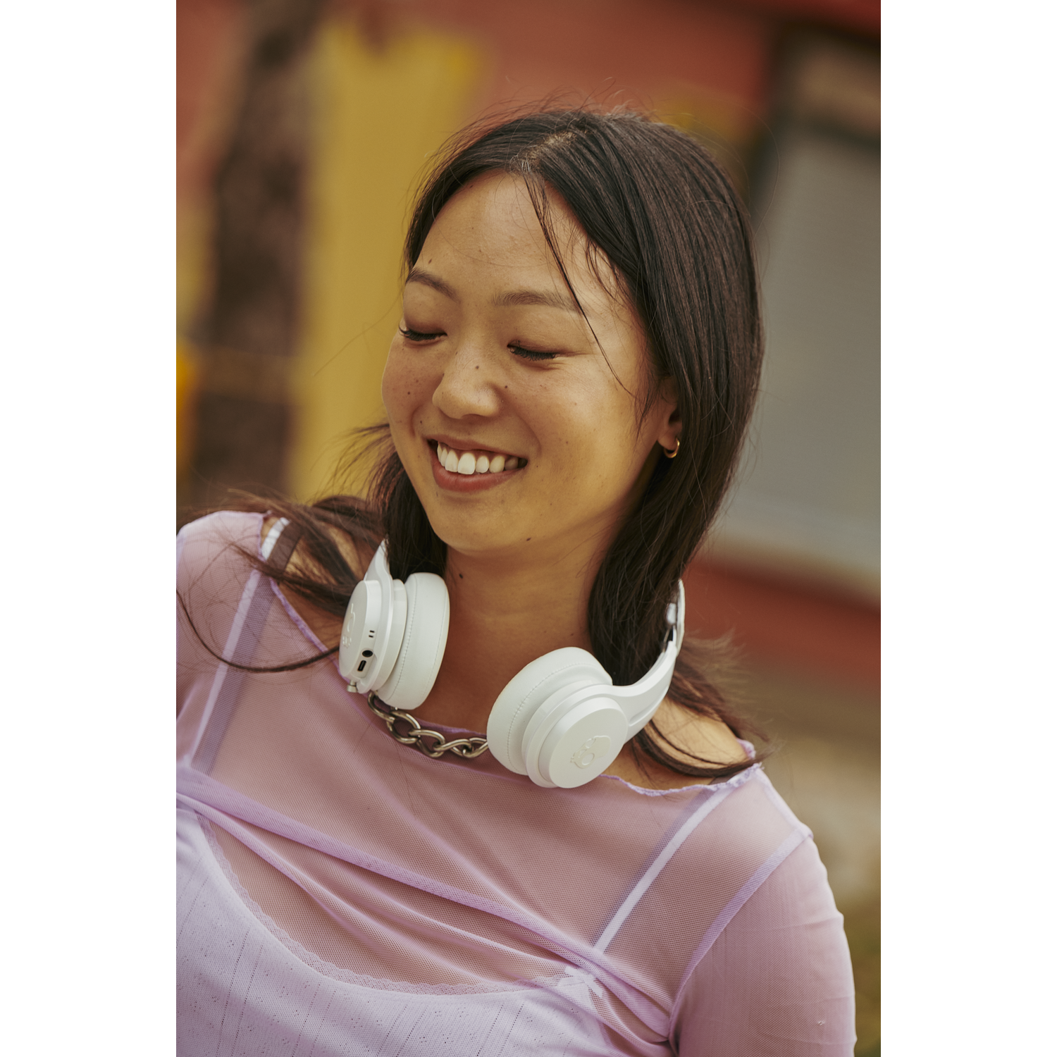 A smiling woman with dark hair wears white headphones around her neck. She is wearing a pink top. Background has color.