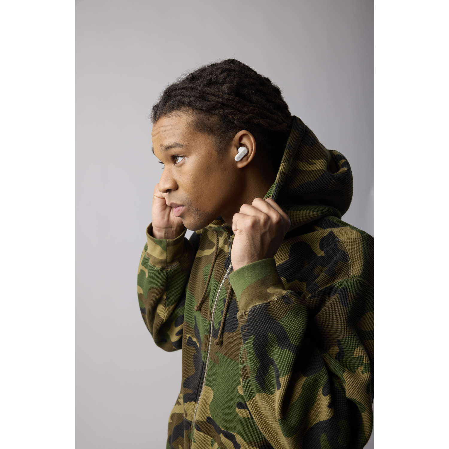A person wearing a camo hoodie wearing white earbuds, looking to the side.