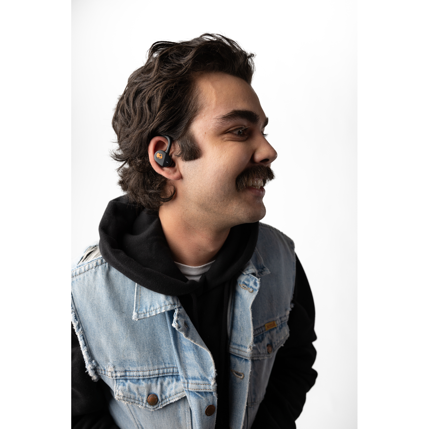 A man with a mustache wearing a denim vest and an earbud in his right ear.