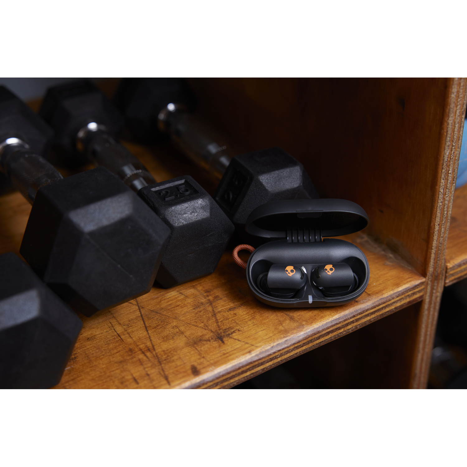 Black earbuds in a case on a wooden shelf. Dumbbells are in the background.