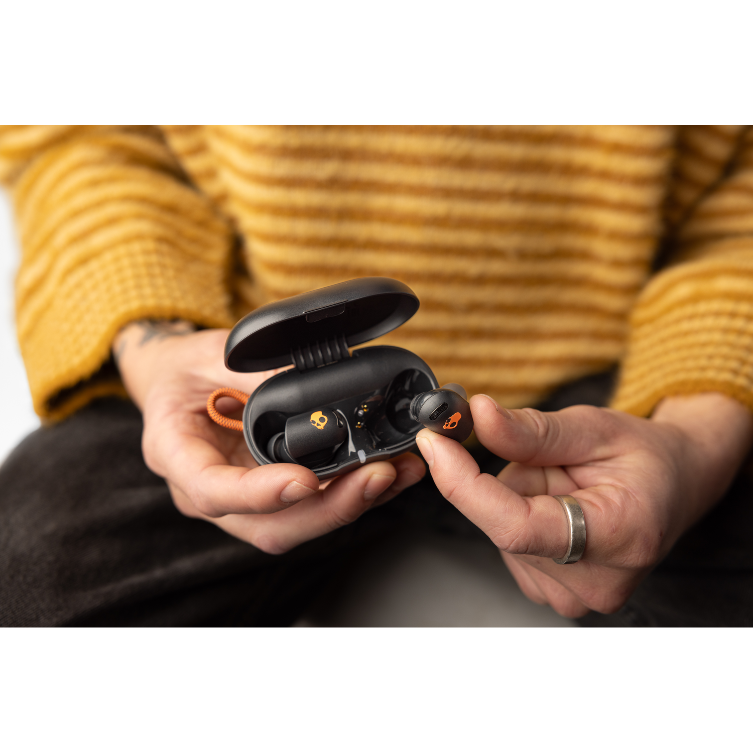 A person is holding a black earbud case and an earbud. The case is open, the setting is indoors.