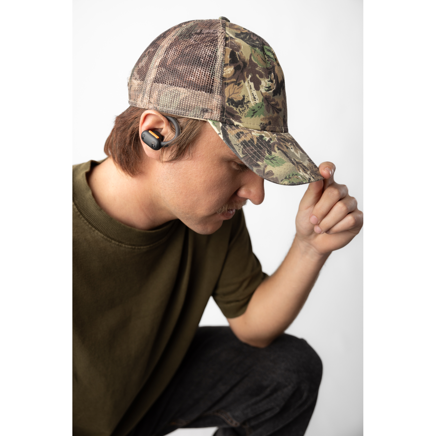 A man wearing a camouflage cap and wireless earbuds, looking down, with a white background.