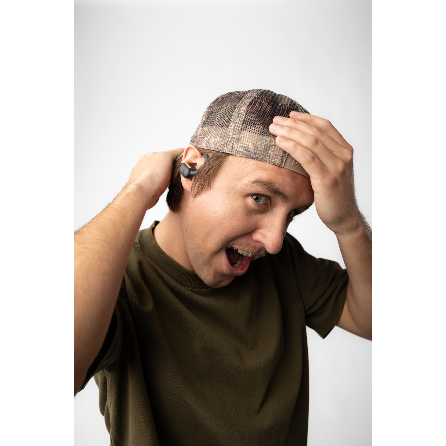 A man wearing a cap and holding his ears, is surprised. He is wearing an olive shirt.