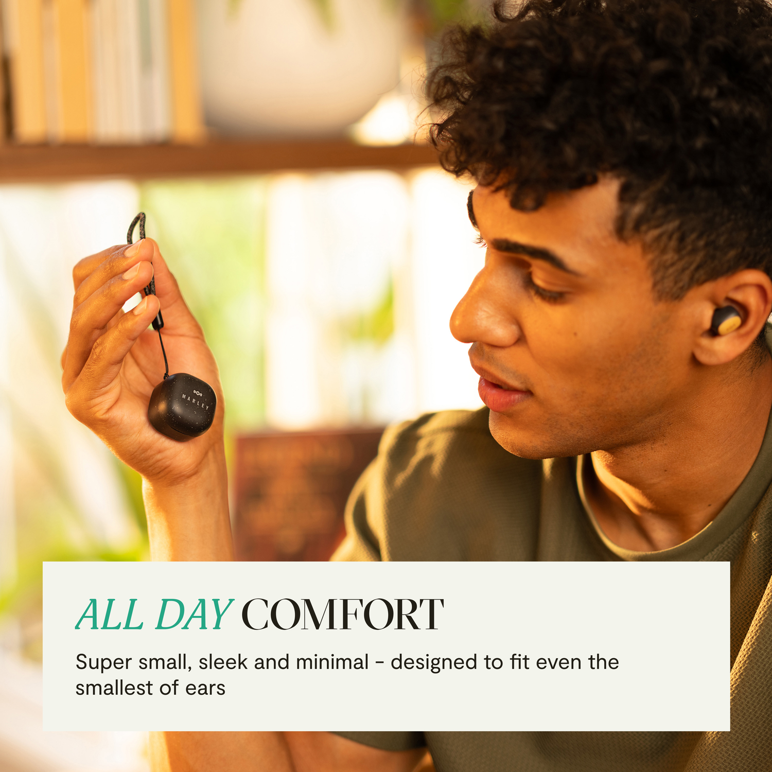 A man holding a black earbud. Text: ALL DAY COMFORT. Background: a room with a window.