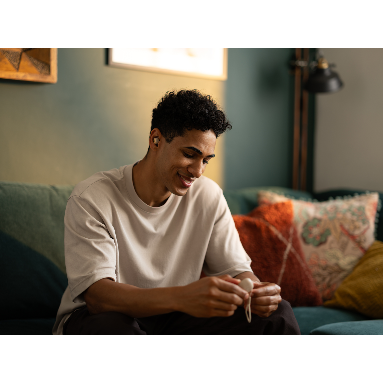 Man sitting on a couch with earbuds in, smiling.  Background includes a painting and a lamp.