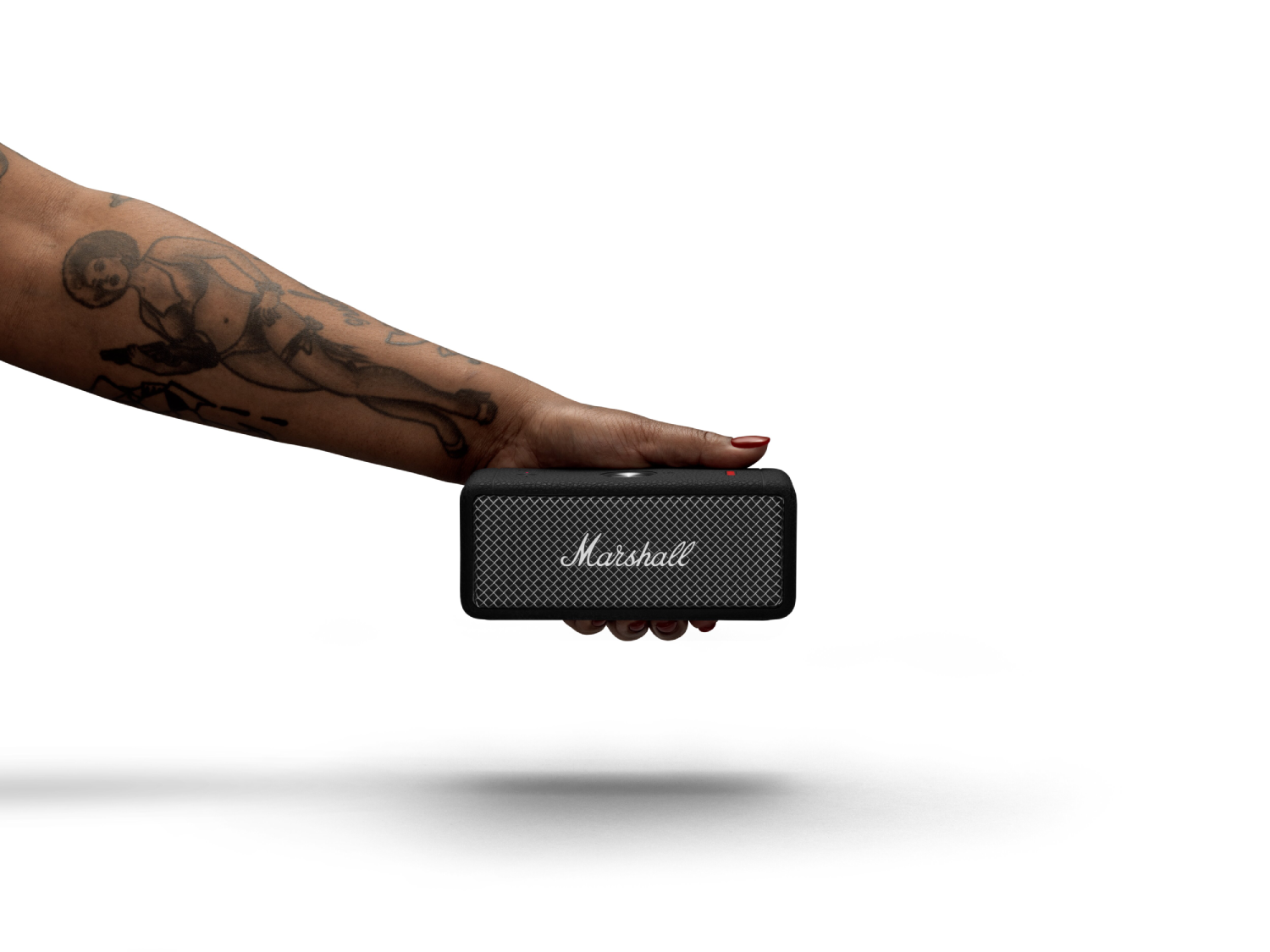 Brazo de una persona con tatuajes sosteniendo un pequeño altavoz Marshall negro. Fondo blanco.