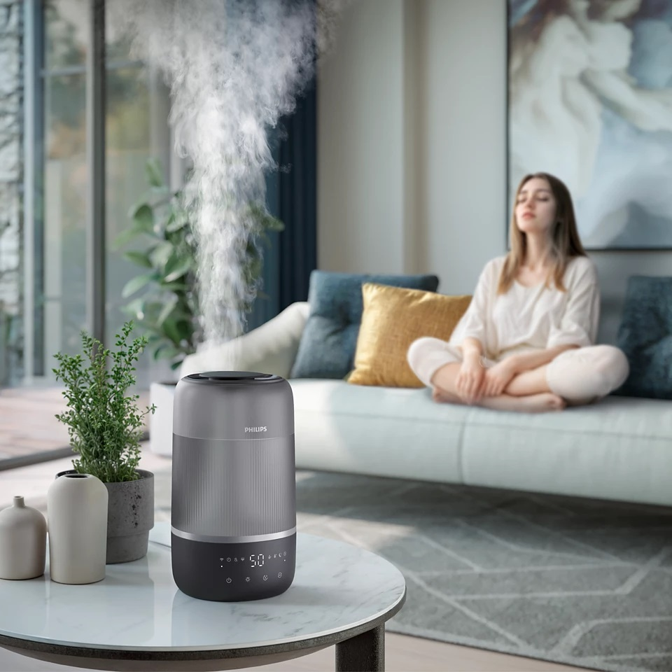 Nawilżacz powietrza w pokoju z kobietą. Nawilżacz jest szary i czarny. Kobieta siedzi na kanapie.