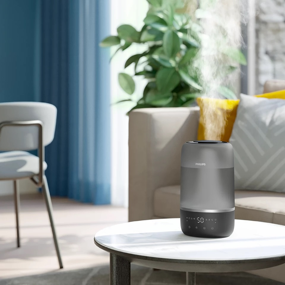 Nawilżacz powietrza na stole. Jest szary i ma wyświetlacz cyfrowy. W tle kanapa i rośliny.