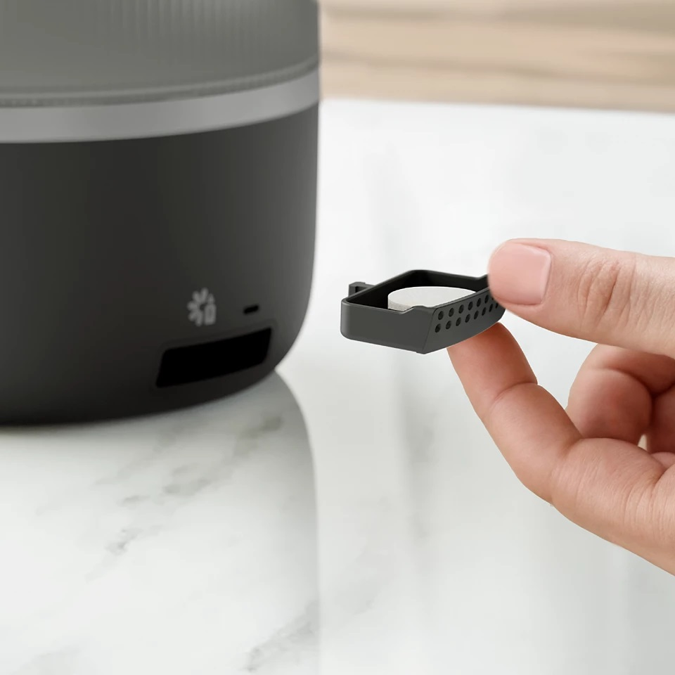 Dłoń trzymająca mały czarny filtr obok ciemnoszarego urządzenia na białej powierzchni.