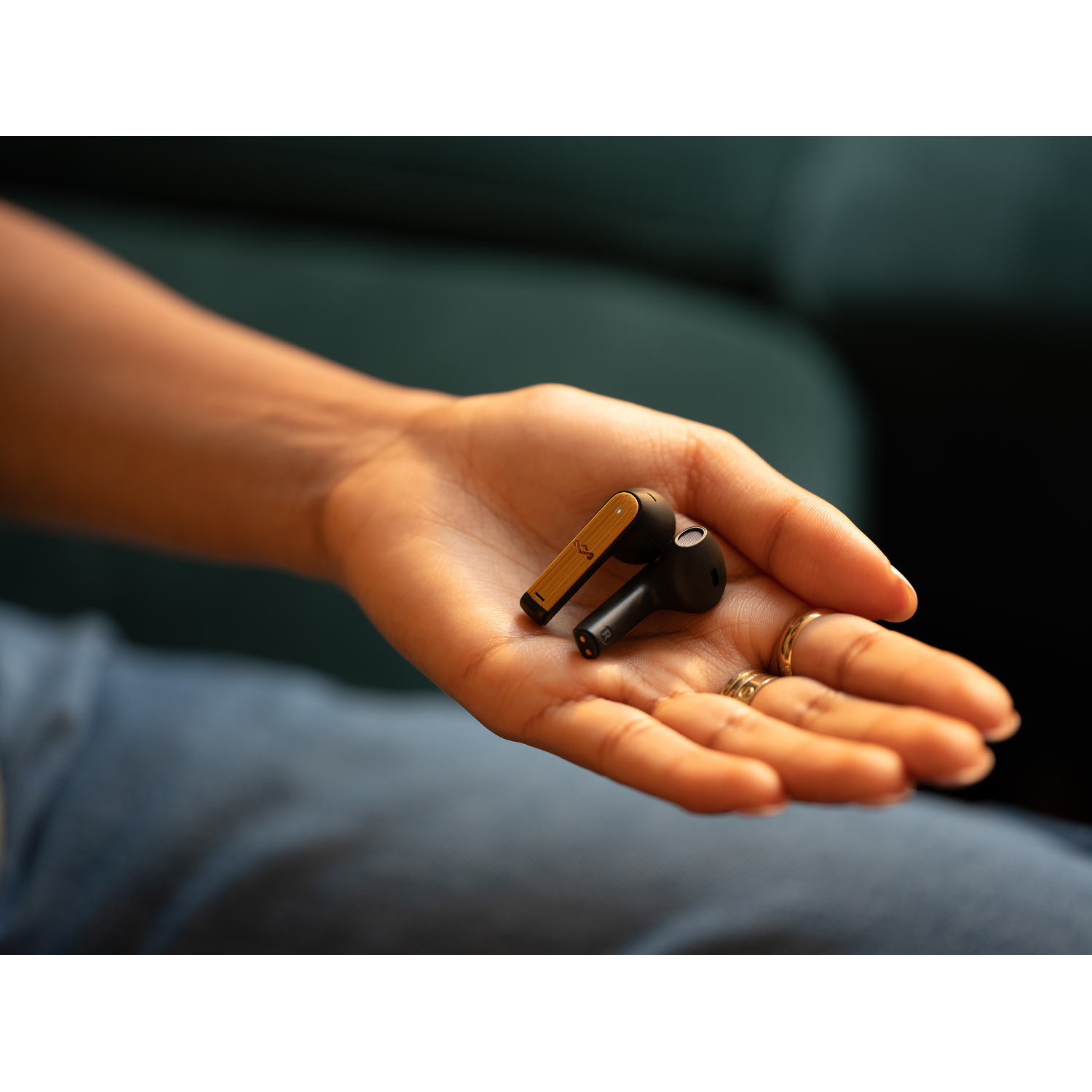 A hand holding two black and gold wireless earbuds. The person is wearing rings.