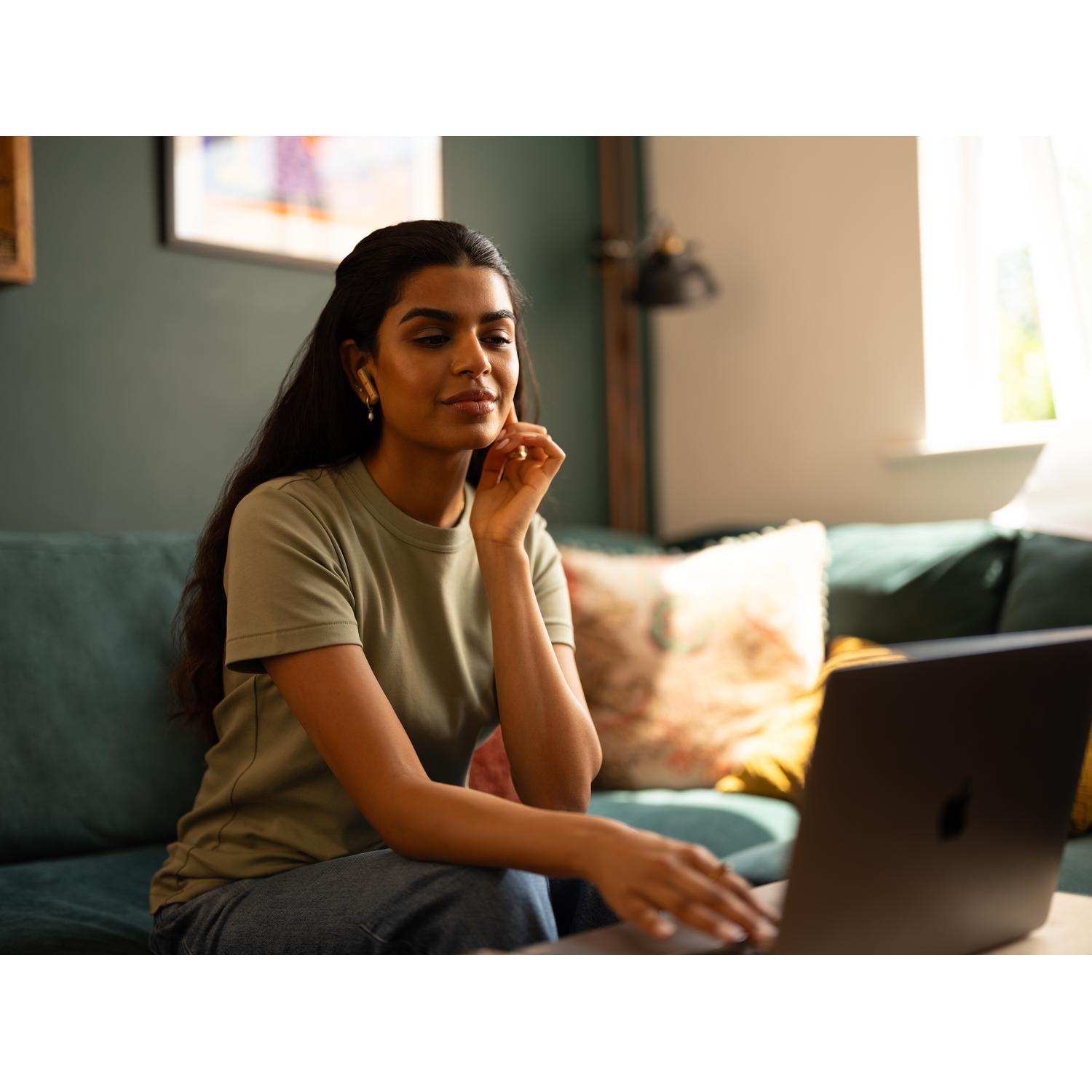 Vrouw zit op een teal sofa, werkt op een laptop. Ze draagt een olijfgroen shirt en gouden oorbellen.