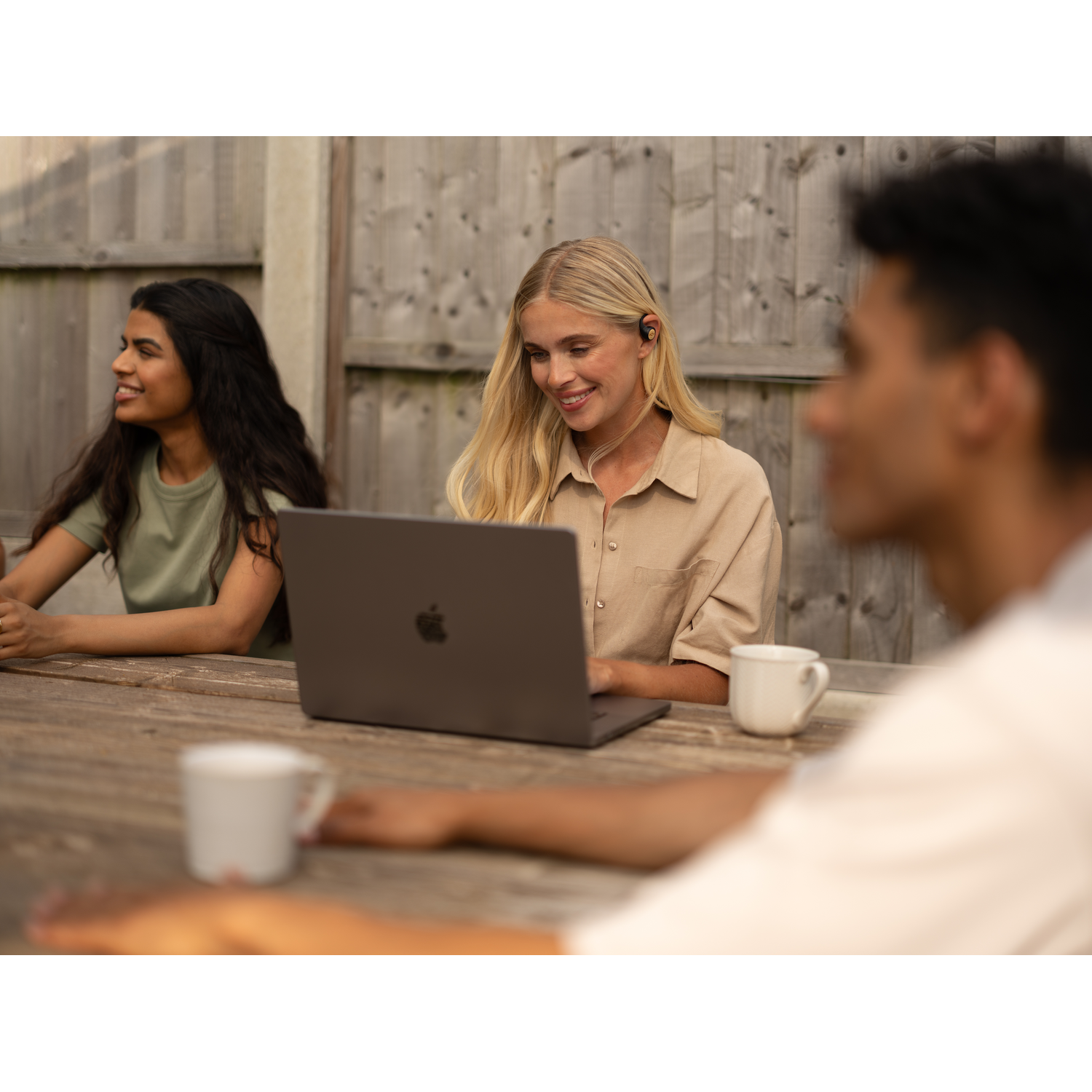Three people with headphones, one using a laptop outside. Wooden table.