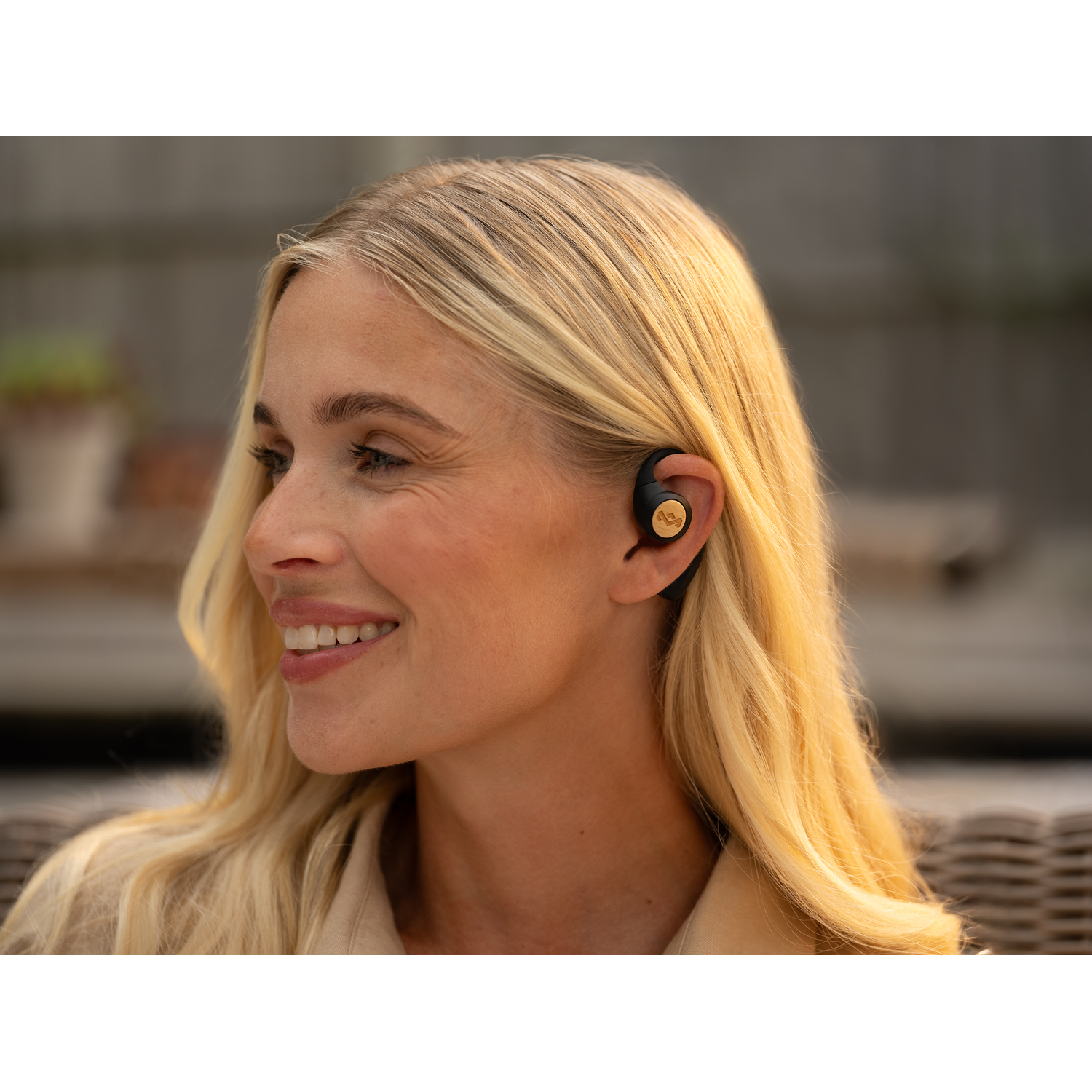 Blonde woman smiling, wearing an earbud, outdoor setting, side view.