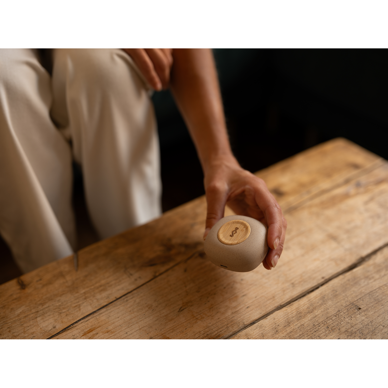 A hand holding a small, round, beige speaker, on a wooden table.