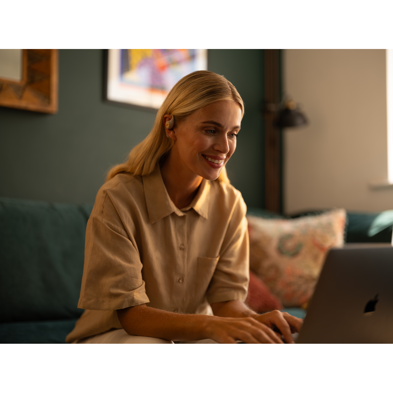 Blond woman working on a laptop. She is smiling and wearing earbuds. Background is a living room.