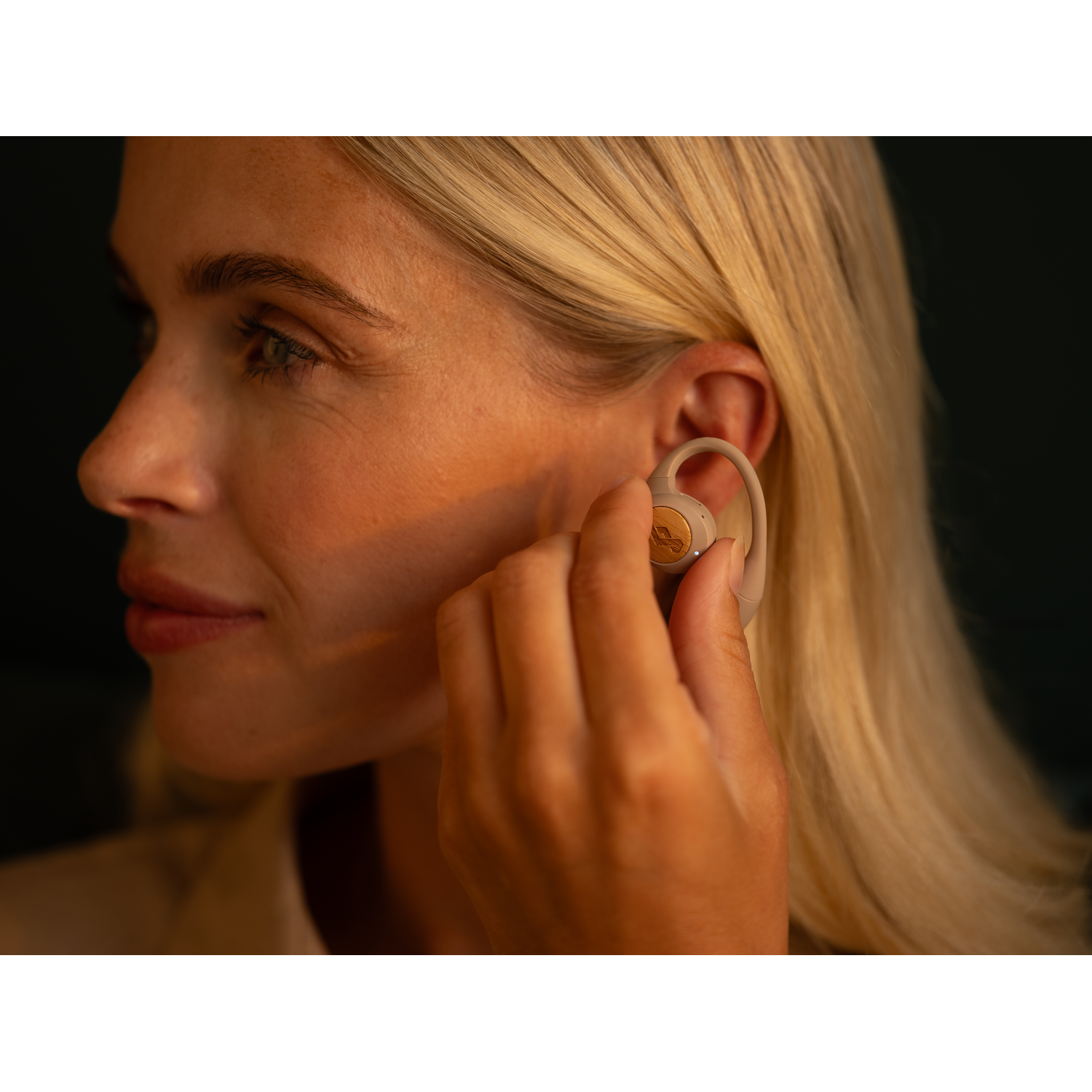 A woman wearing a beige earbud in her ear. She is blonde and holding the earbud.