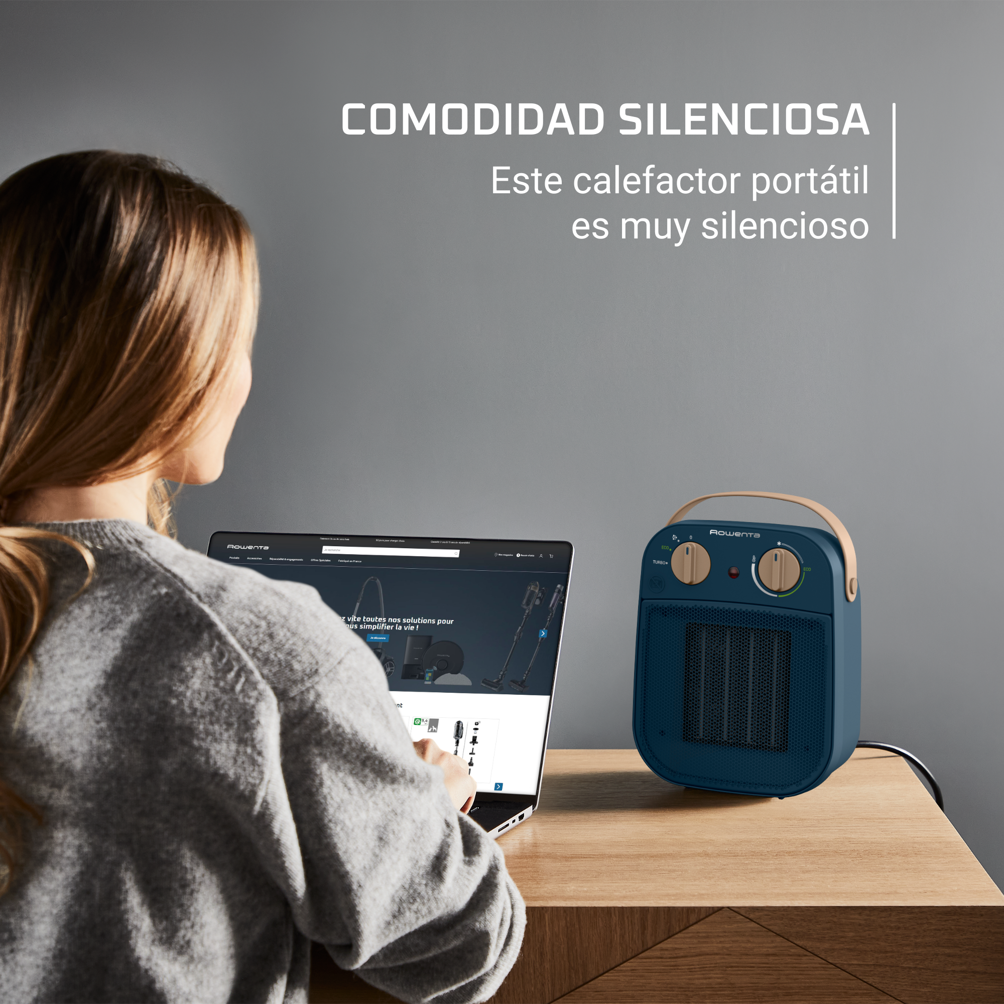 Mujer sentada cerca de un pequeño calentador azul en una mesa de madera, usando una laptop.