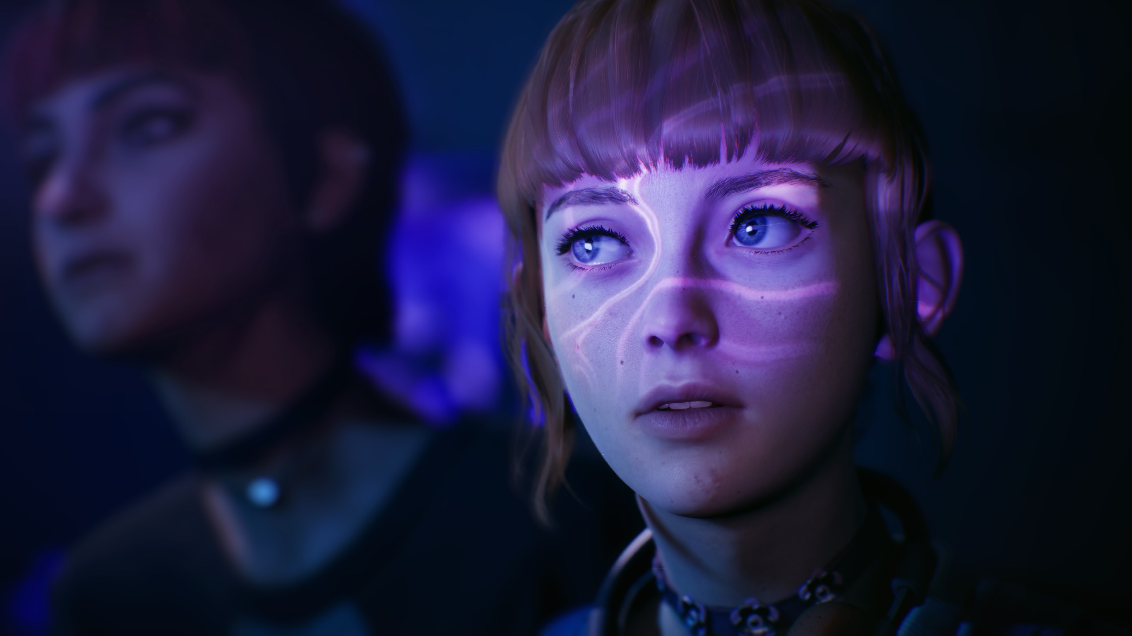Una donna con occhi blu e capelli rossi guarda in alto. Motivi di luce viola sul suo viso. Un'altra persona è sfocata sullo sfondo.