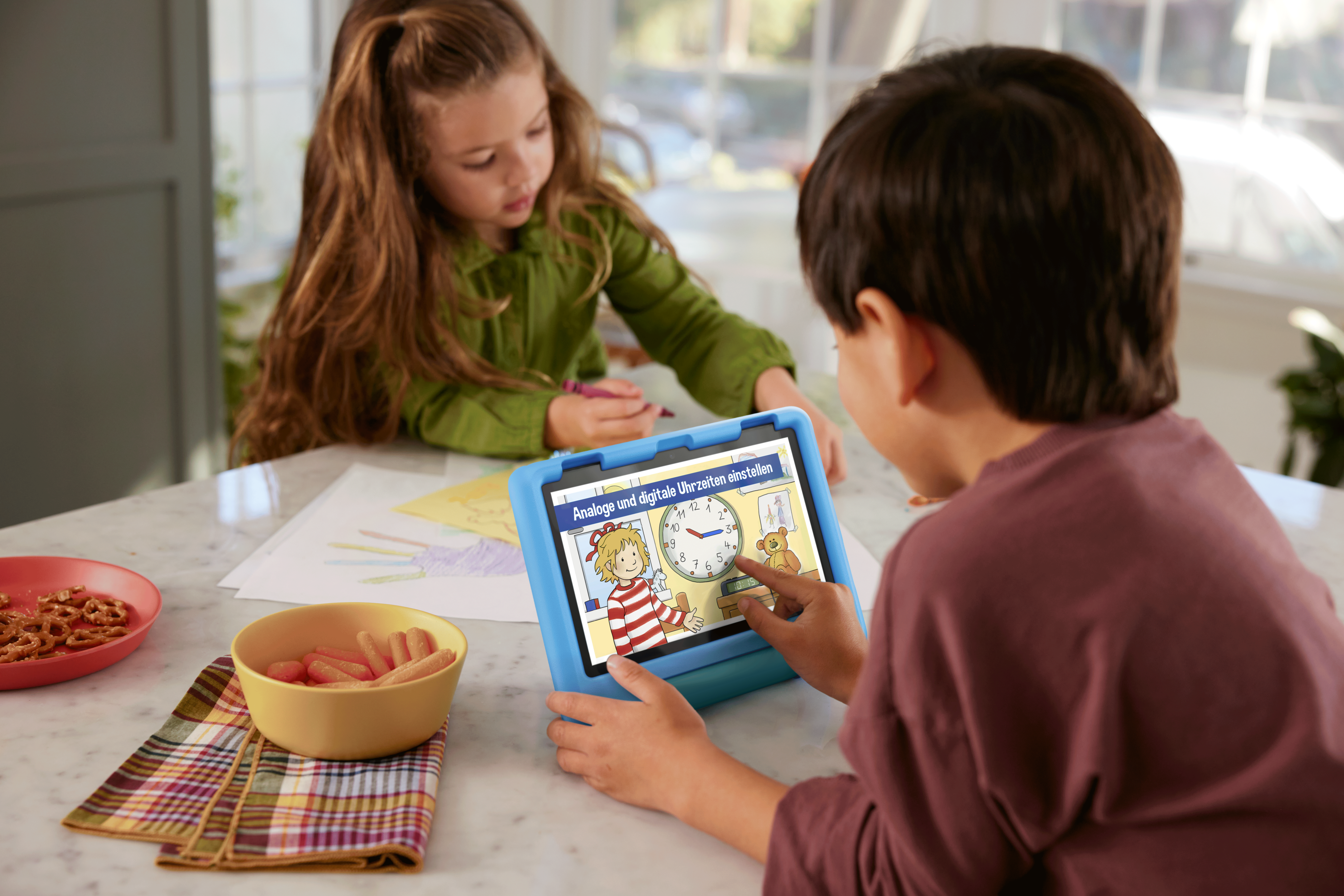 Zwei Kinder schauen sich ein Tablet an. Das Tablet zeigt eine Uhr. Auf dem Tisch liegen Snacks.