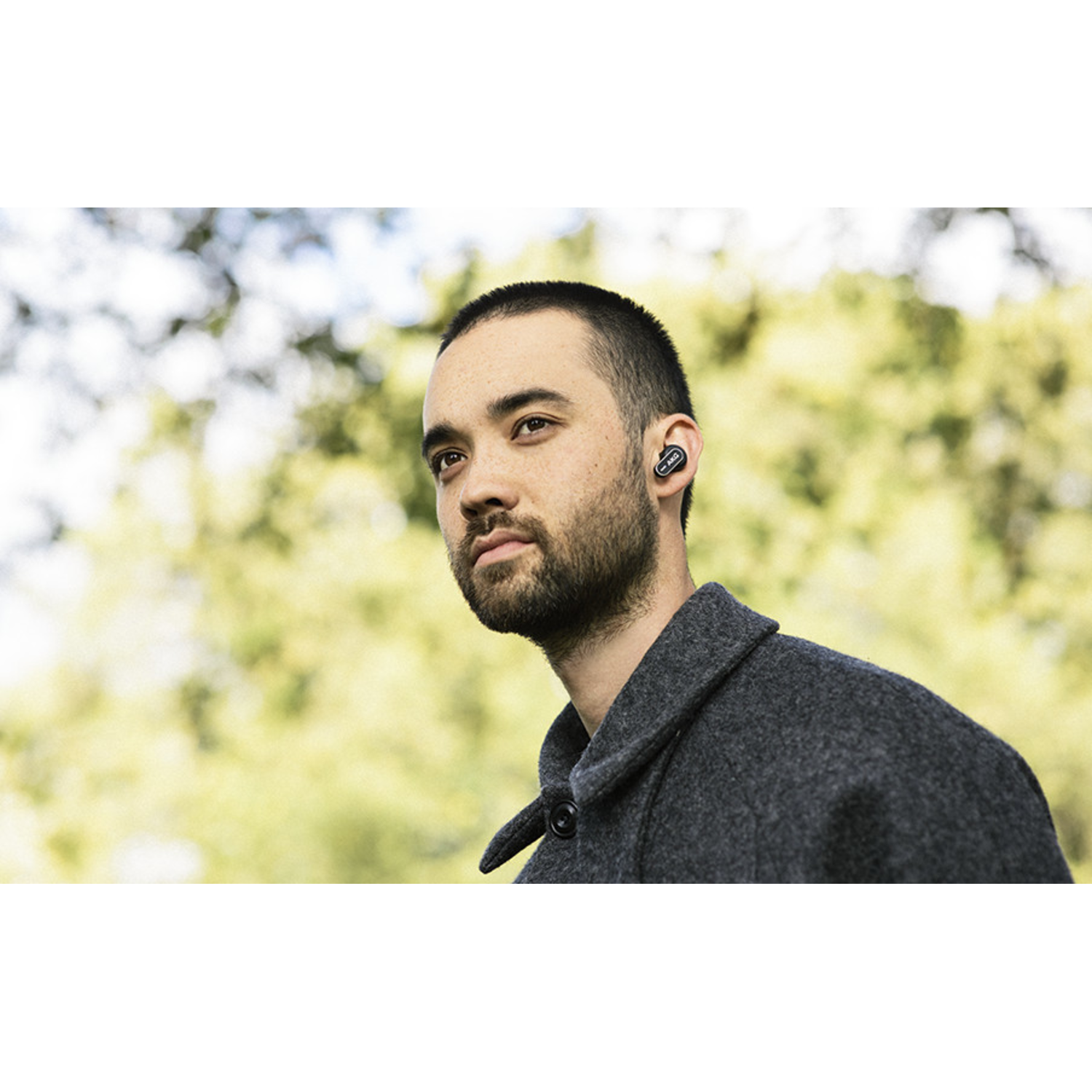 A man with a beard wearing an earbud in his ear. The background is a blurred view of trees.