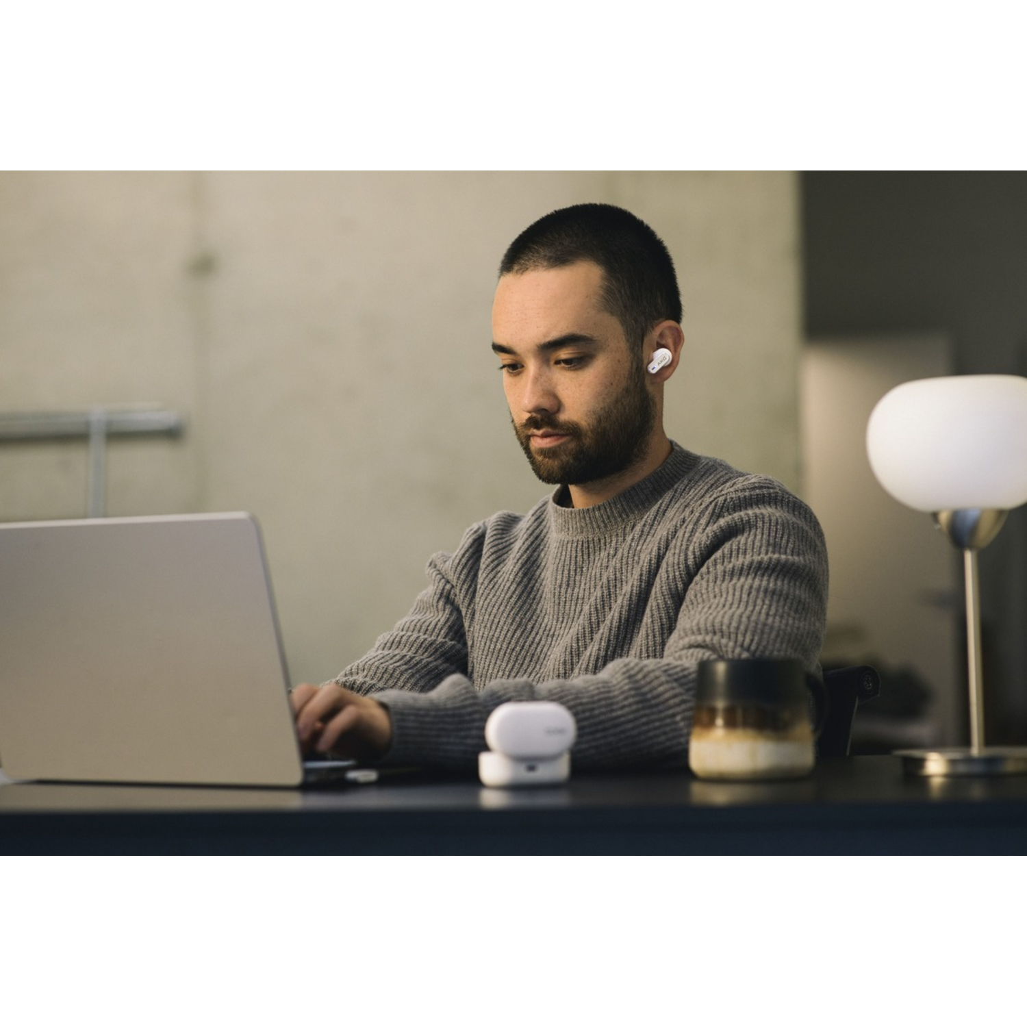 Man wearing earbuds, working on a laptop at a desk with a lamp and a coffee cup.