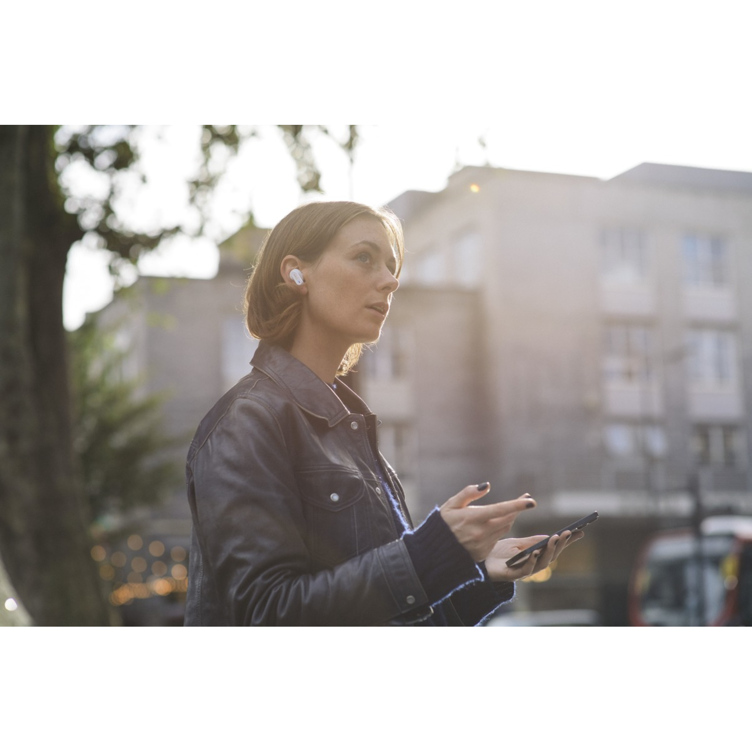 Woman wearing earbuds, holding phone, gesturing with hands, with buildings in background.