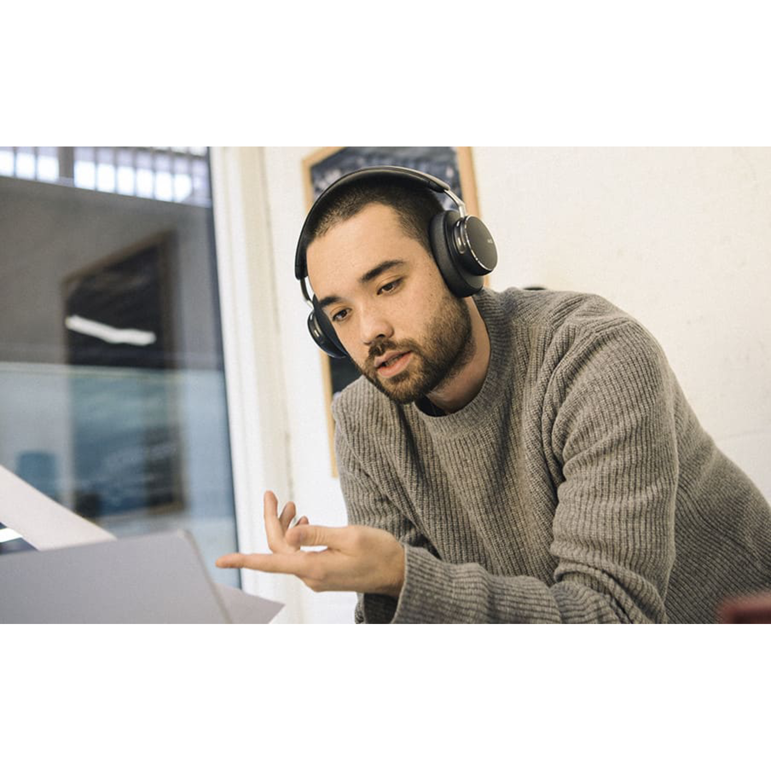 A man wearing headphones looks at a laptop, gesturing with his hand. The background is blurred.