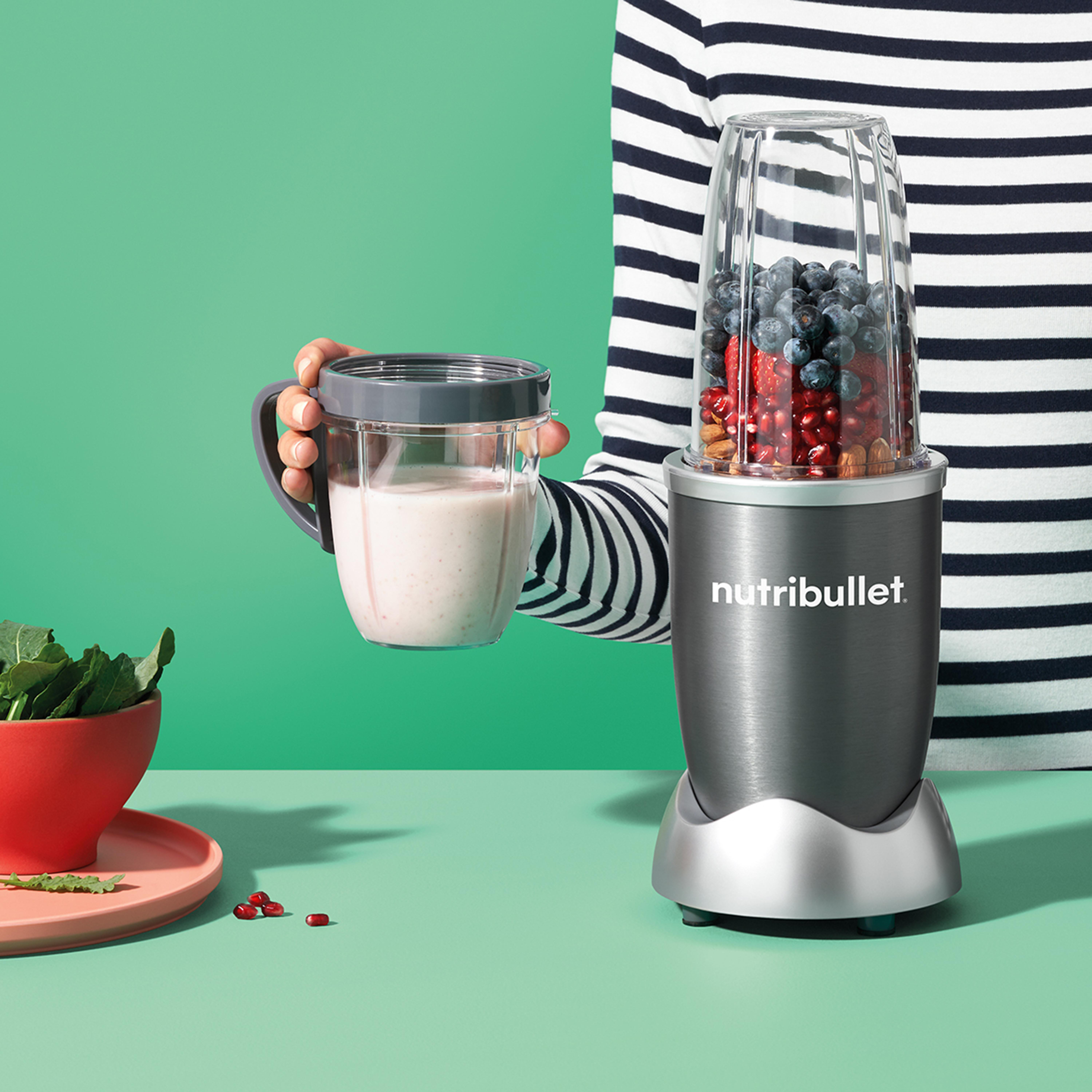 Eine Person hält einen Mixerbecher mit pinkfarbenem Smoothie. Ein Mixer steht daneben, gefüllt mit Zutaten.