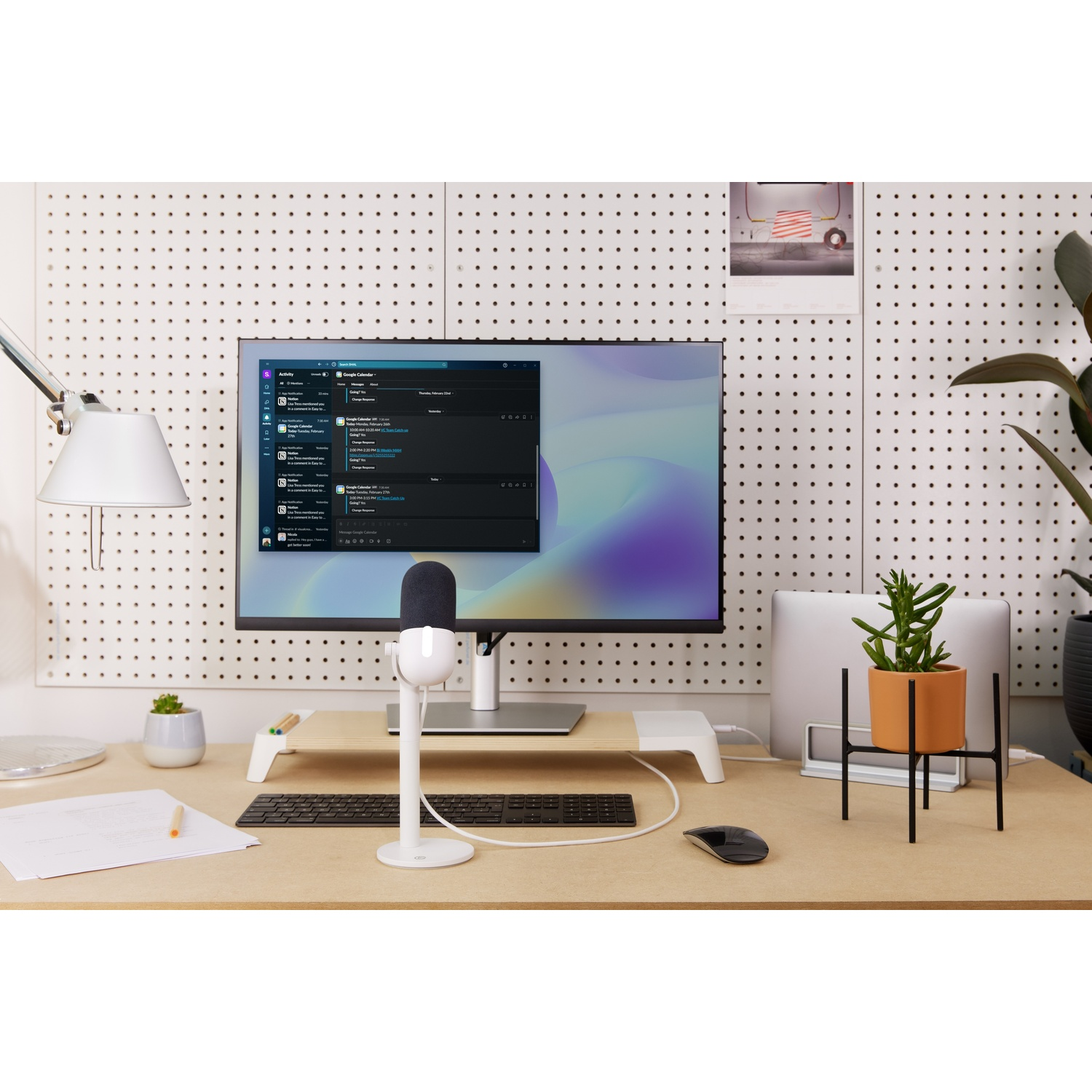 A desk setup with a monitor, microphone, keyboard, and plant. The background is white.