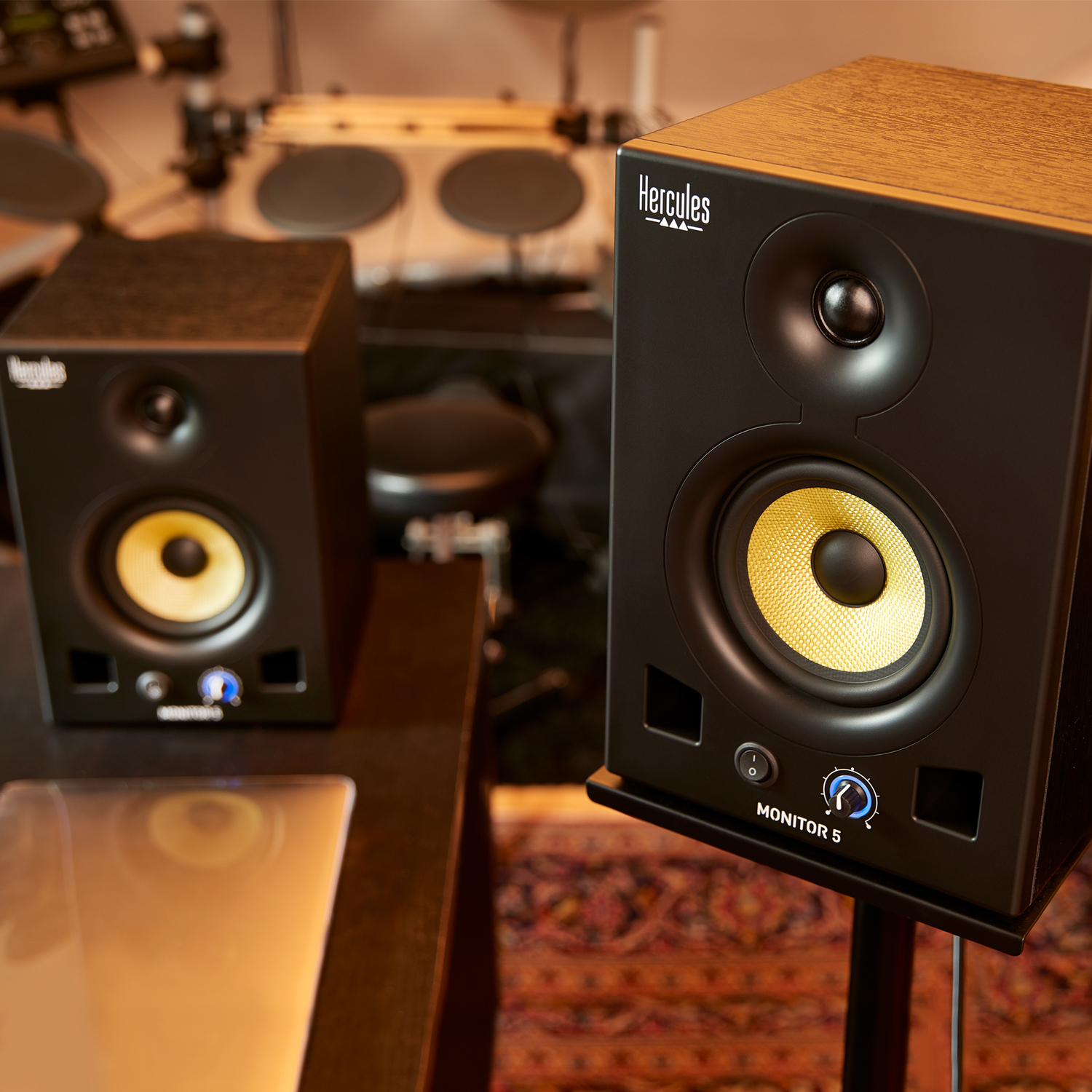 Twee zwarte Hercules Monitor 5 speakers met gele conussen in een studio. Een drumstel op de wazige achtergrond.