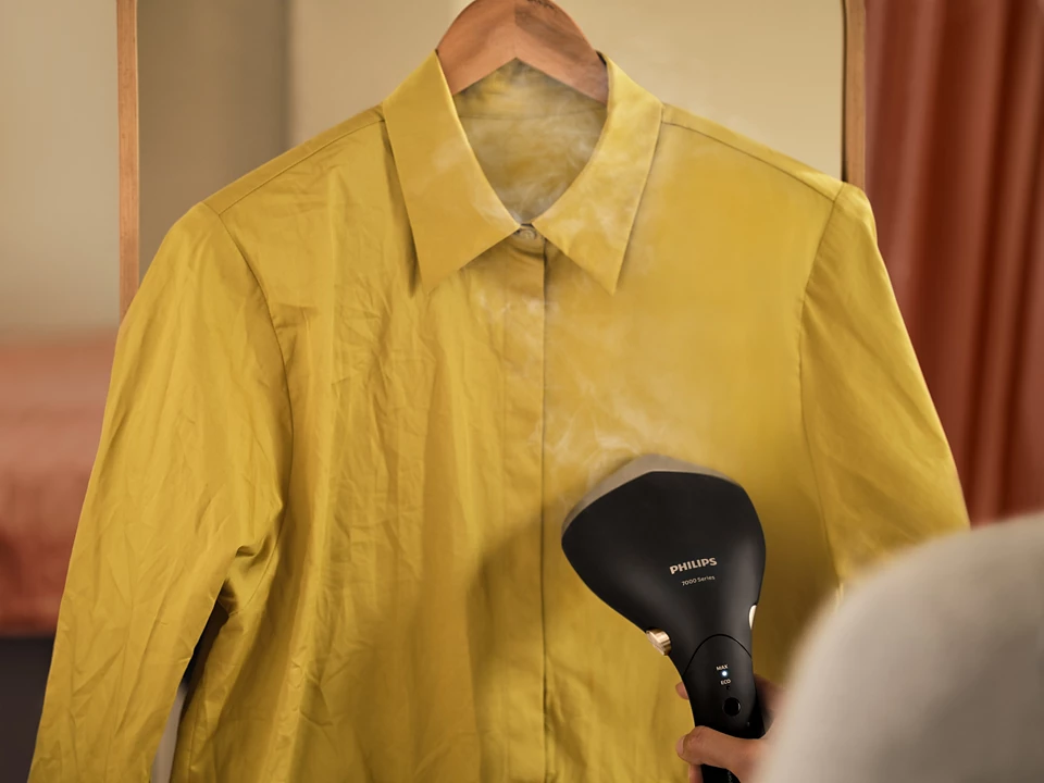 Eine gelbe Jacke auf einem Bügel wird mit einem schwarzen Dampfgarer gedämpft. Ein Spiegel ist dahinter.