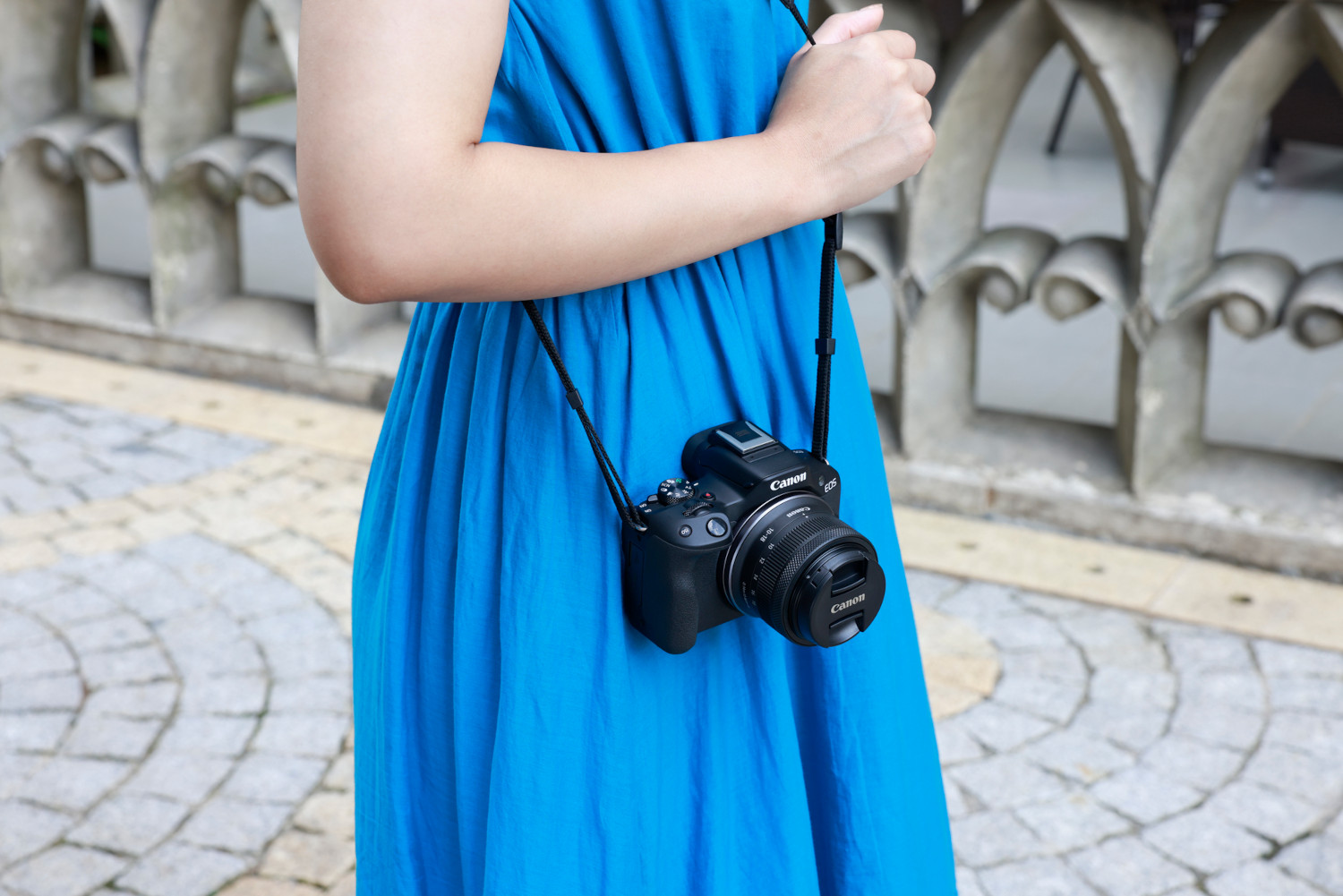 Woman in blue dress holds a black camera. Background is a stone wall.