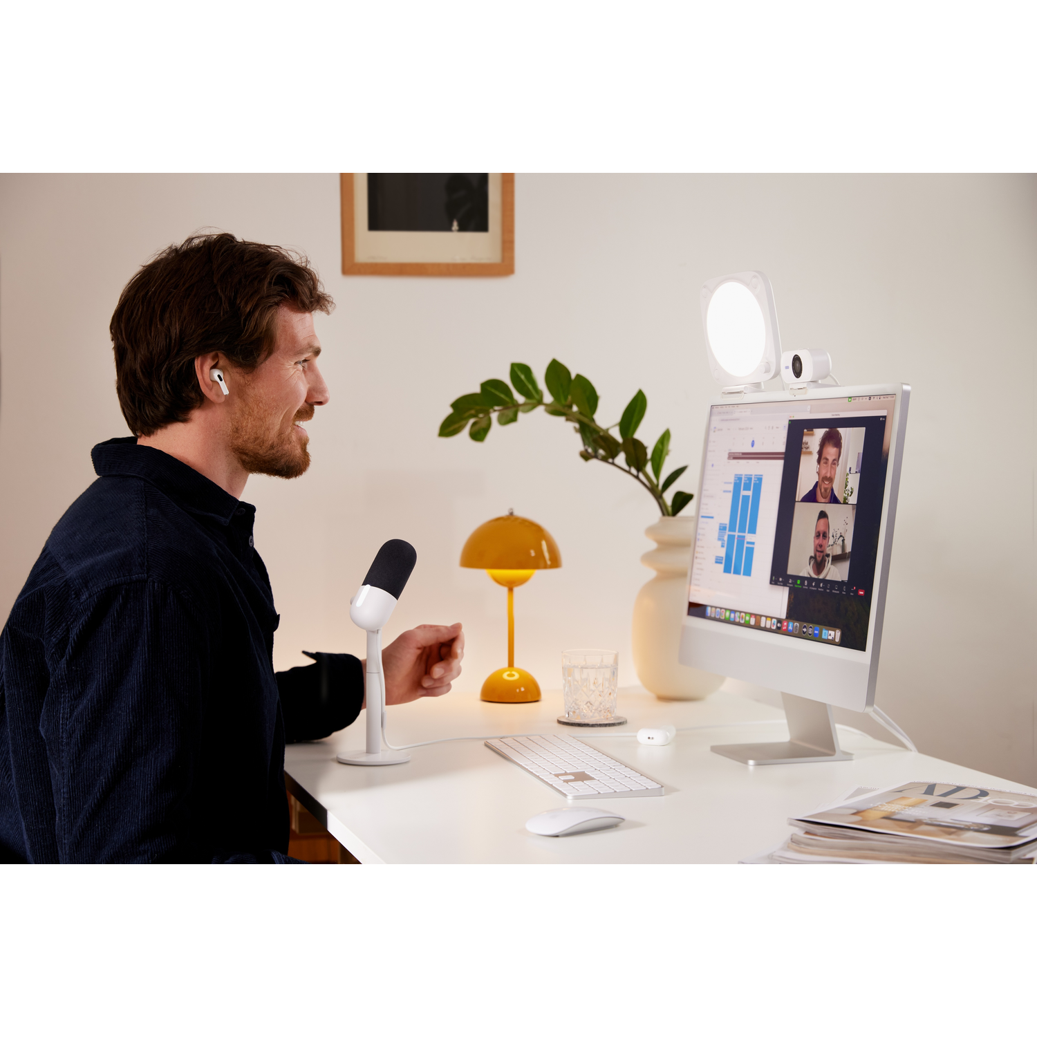 Man in a dark shirt on a video call, computer, microphone, lamp, and a glass are on the desk.