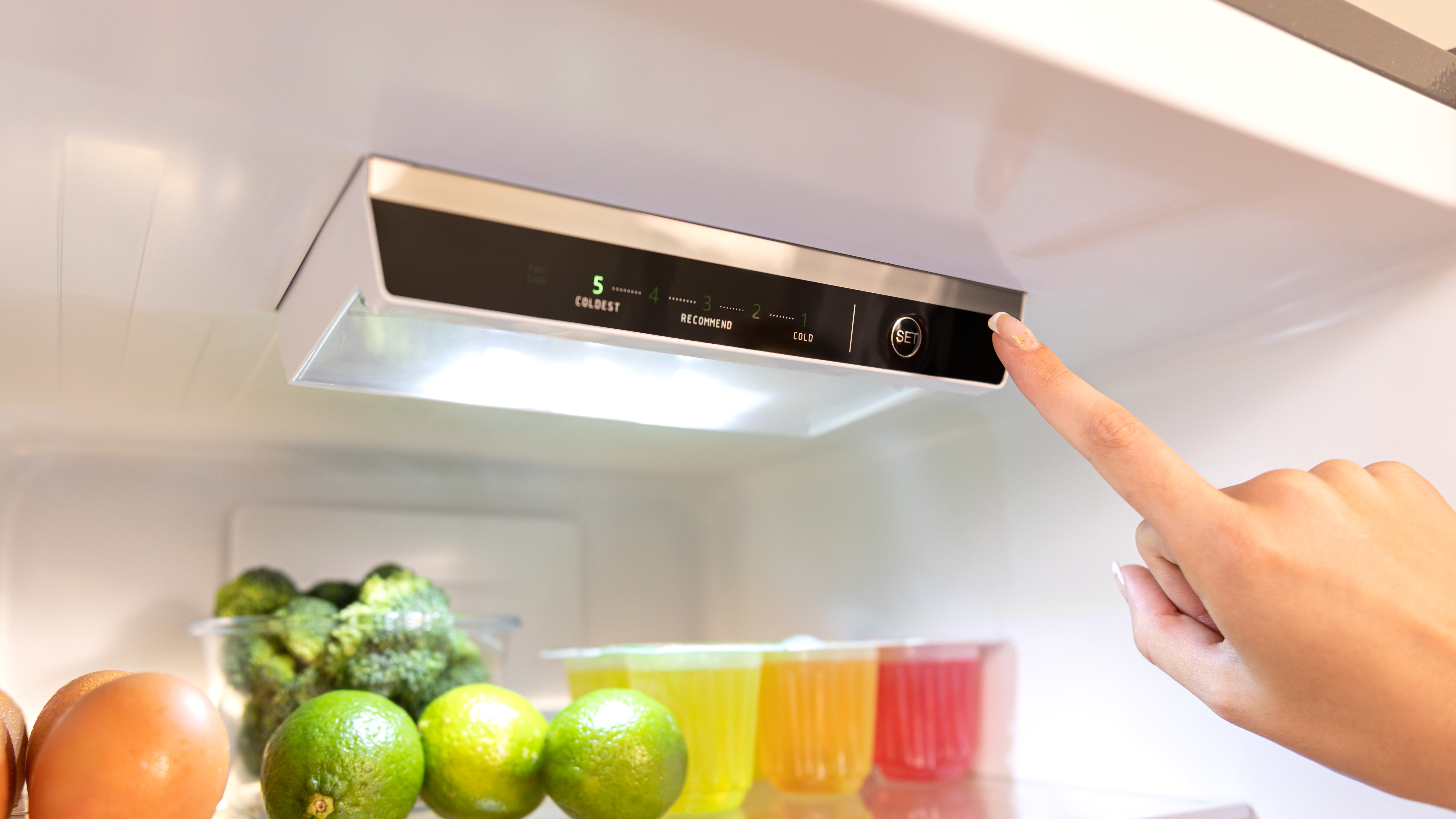 Una mano presiona el botón 'set' dentro de un refrigerador. Frutas y verduras son visibles.