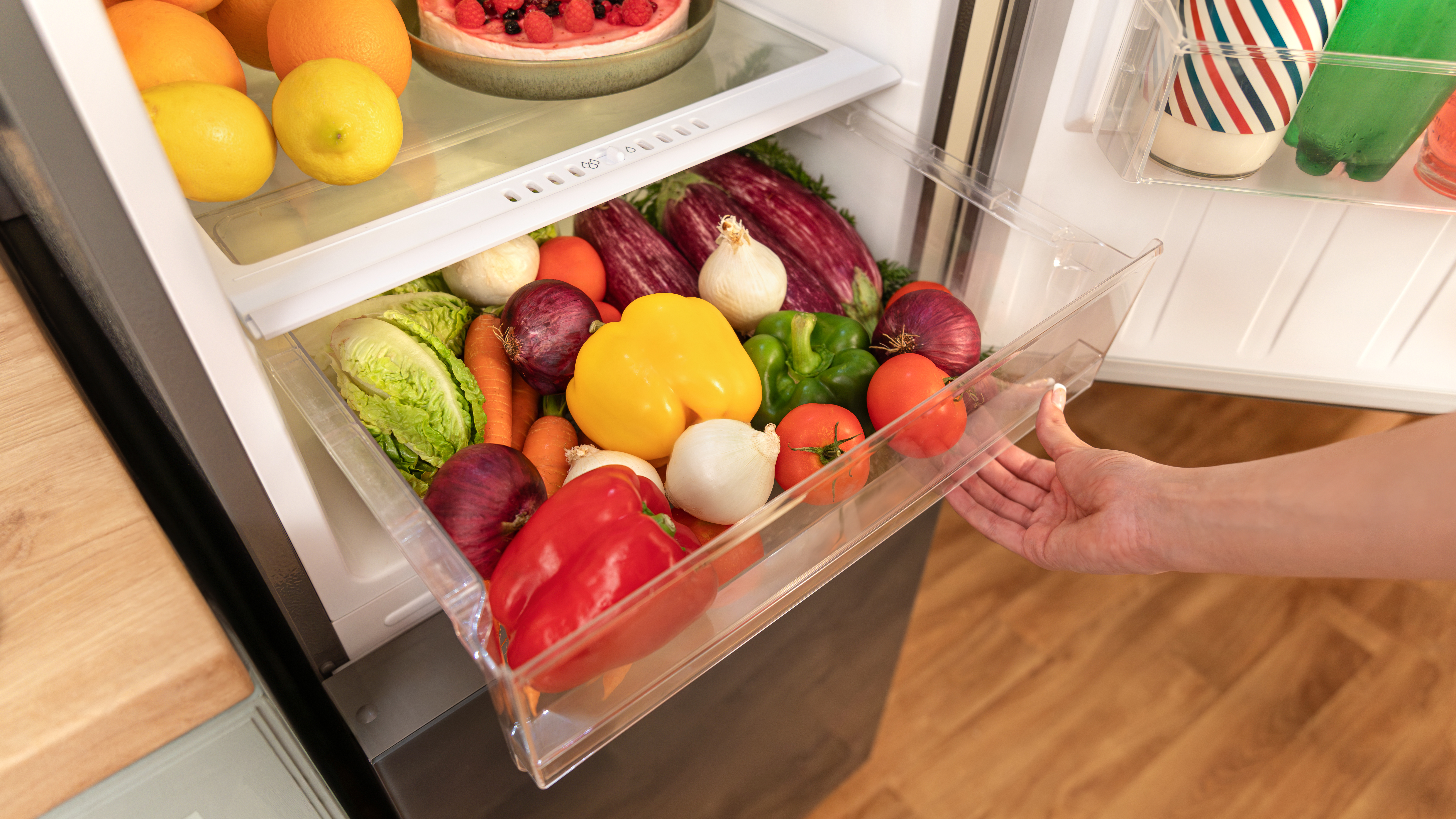 Nevera abierta con cajón de verduras fuera. Incluye zanahorias, pimientos, cebollas y tomates.
